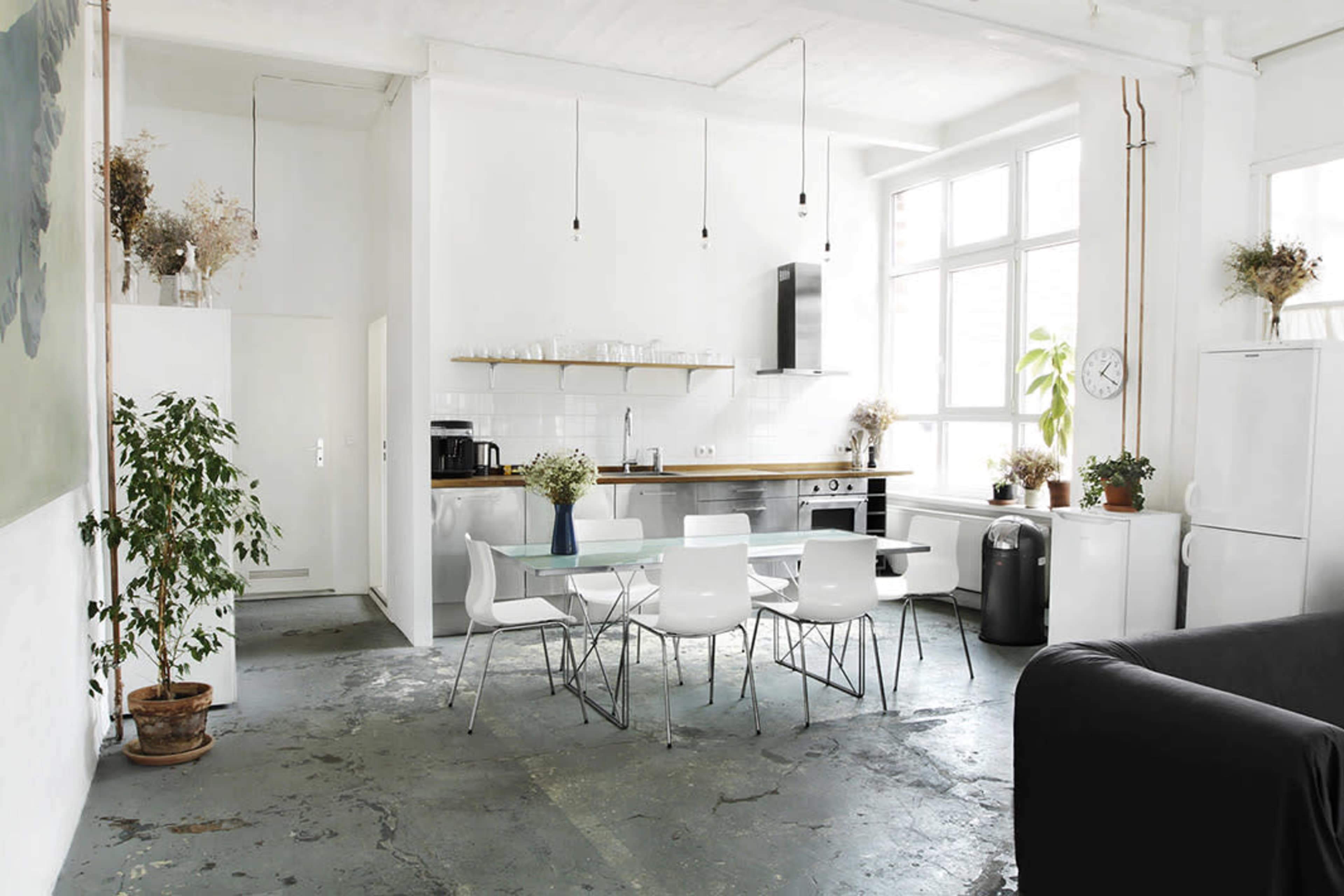 A bright, minimalist kitchen and dining area features a glass table surrounded by white chairs, with plants and a modern appliance setup in a spacious, open layout.