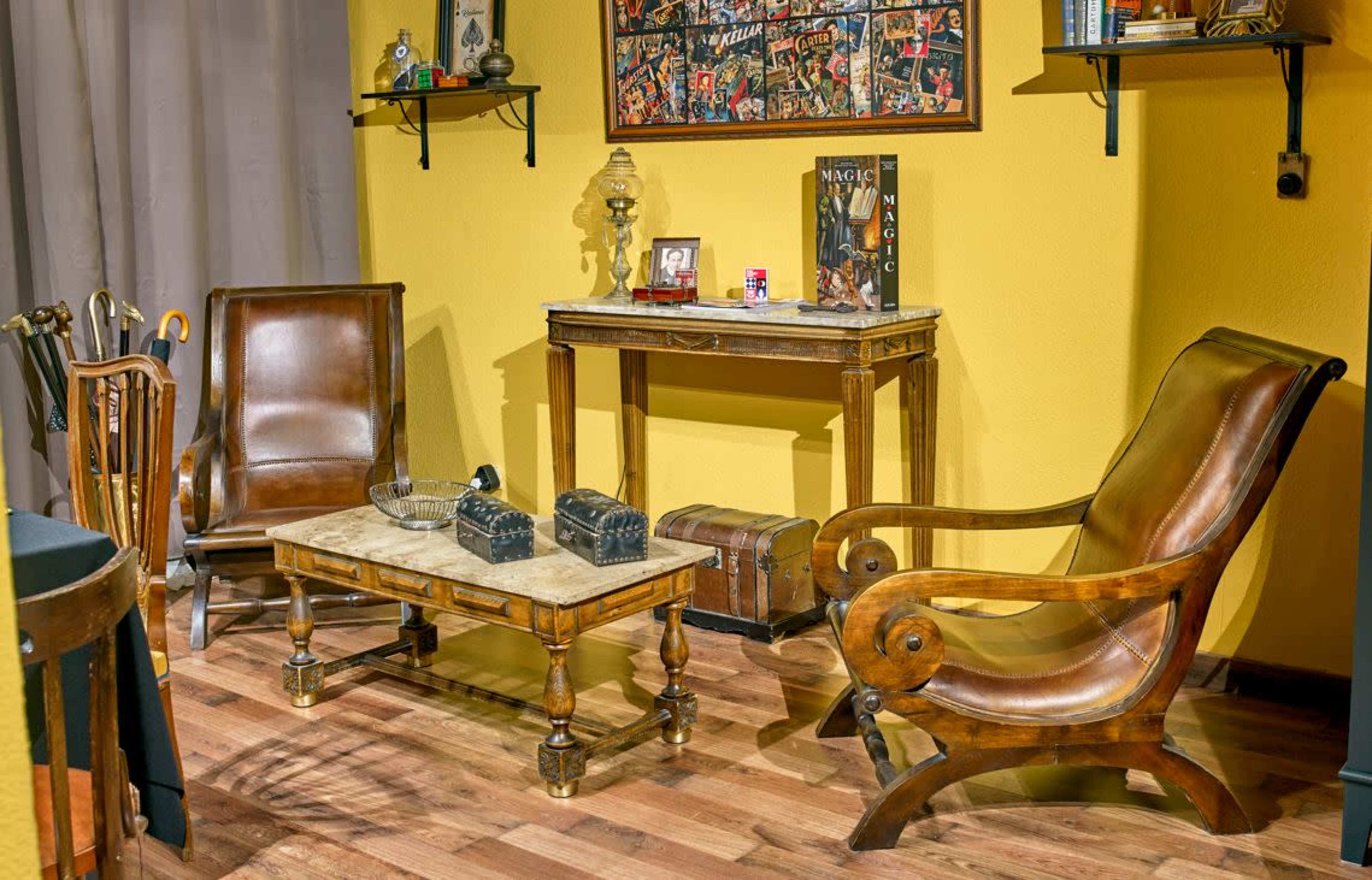 The image shows a cozy room featuring two brown leather armchairs, a marble-topped coffee table, a wooden console table, and a yellow wall decorated with framed photographs.
