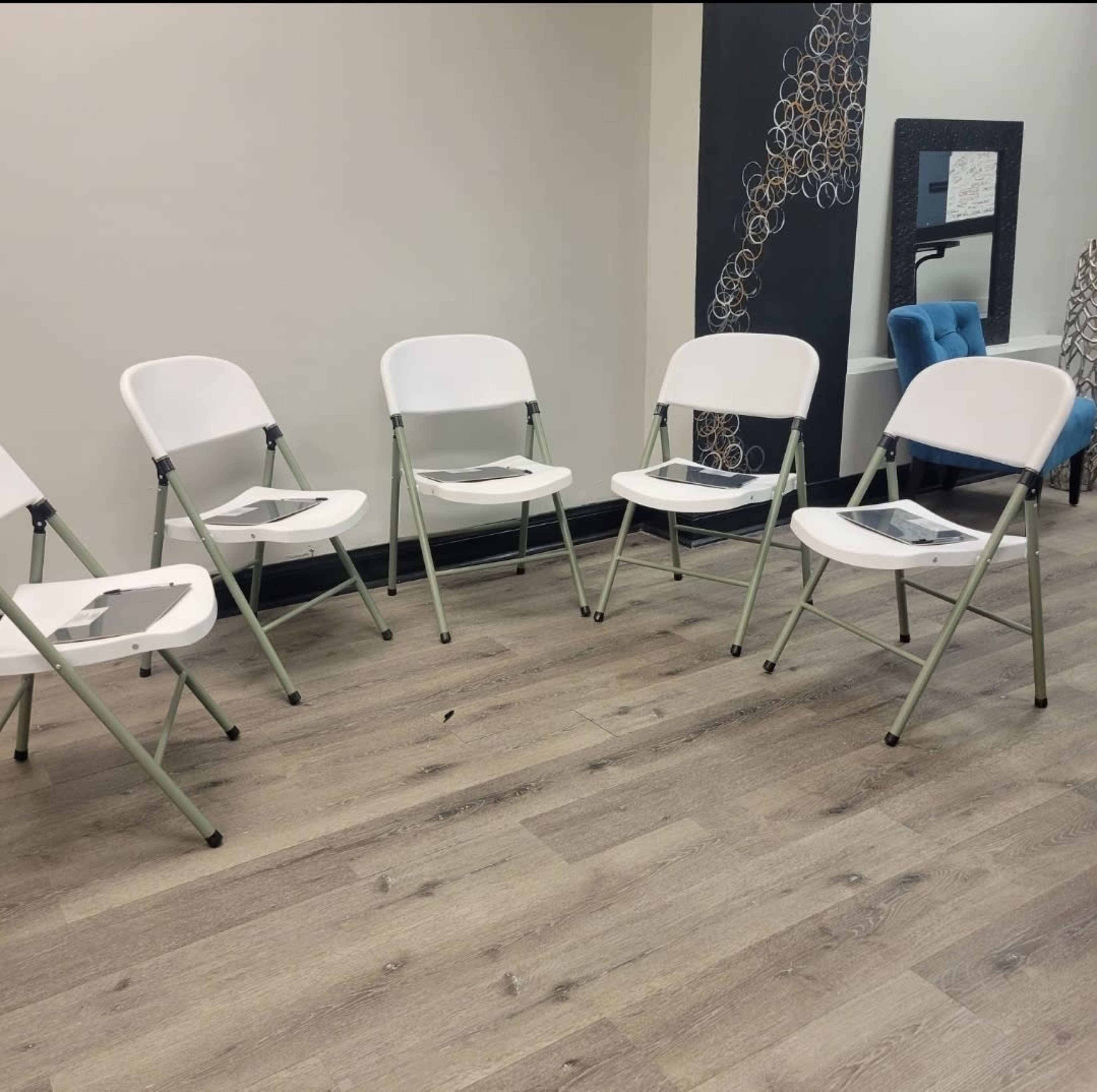 The image shows a row of six white folding chairs arranged on a wooden floor in a minimally decorated room.