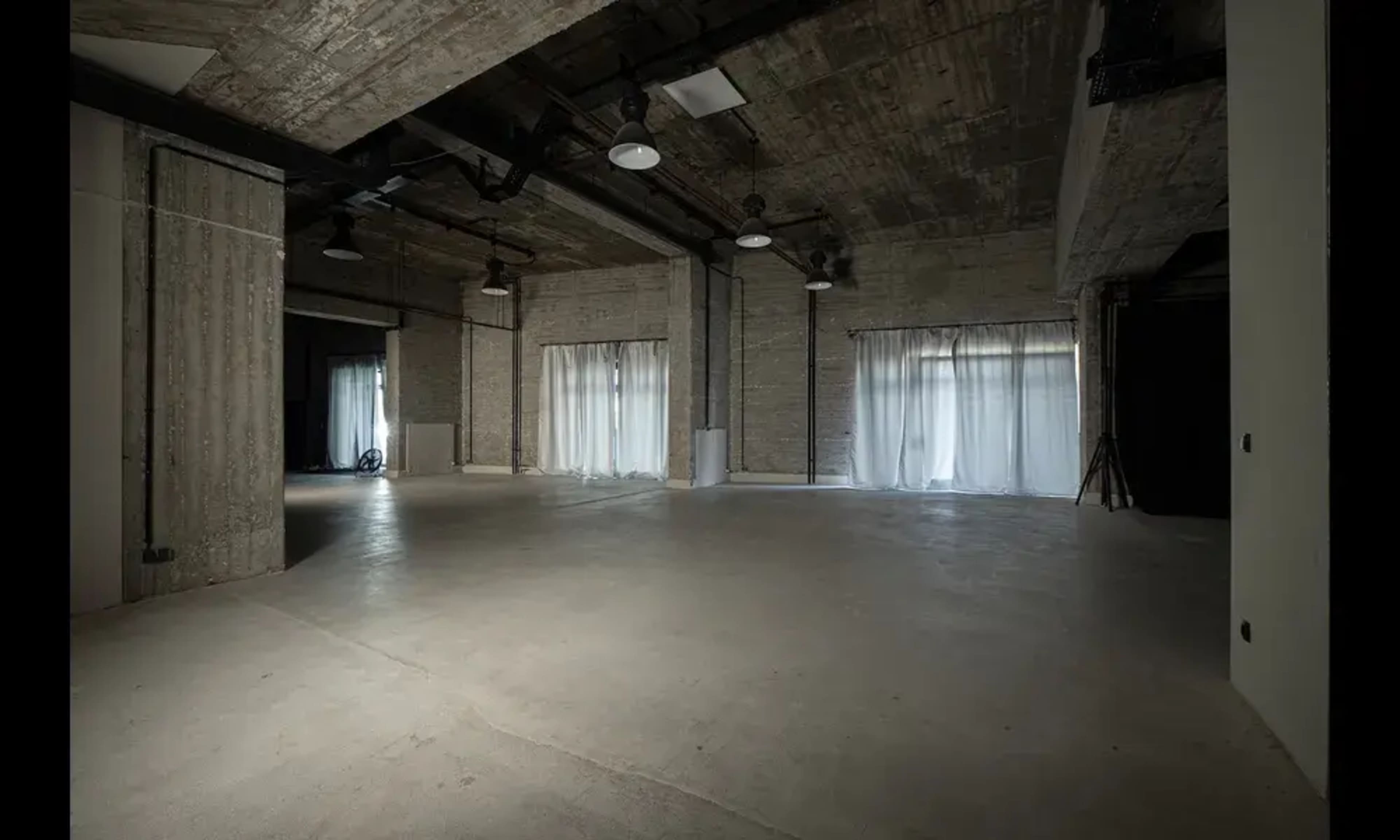 The image shows a spacious, industrial-style room with concrete walls, large windows covered by sheer curtains, and a polished concrete floor.