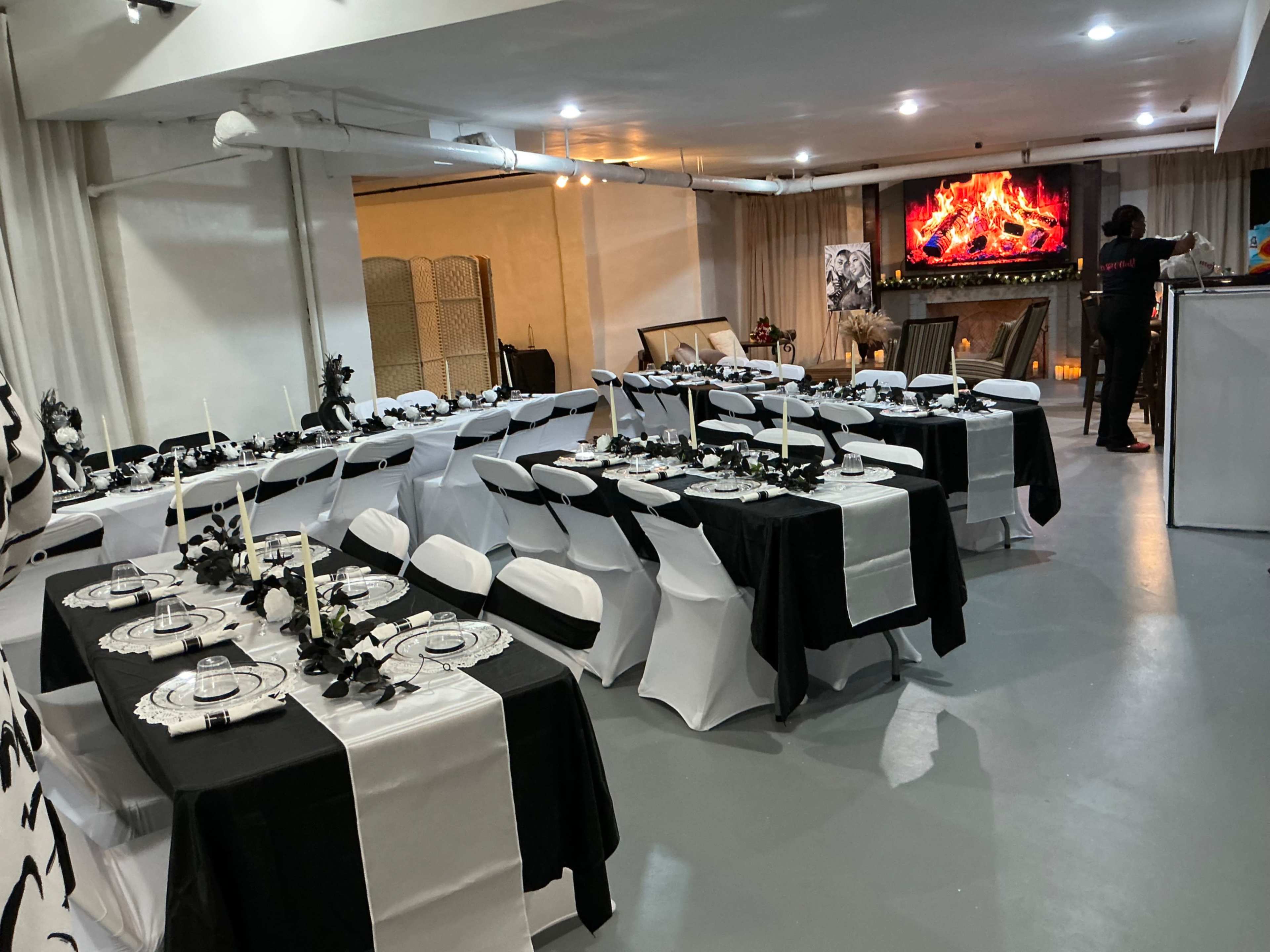 The image shows a banquet hall set for a dinner with black and white table settings, candles, and a large screen displaying a digital fireplace in the background.