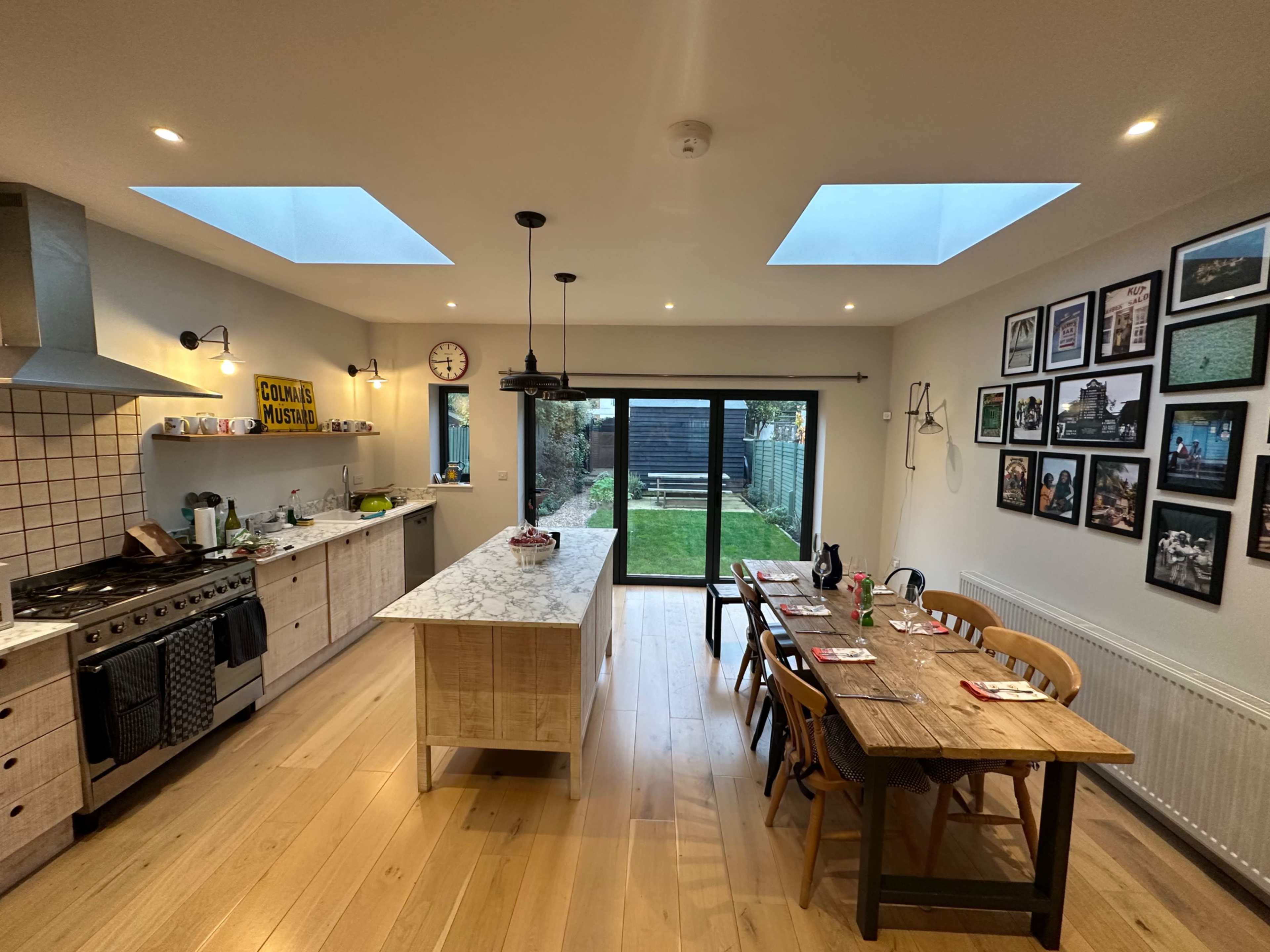 A modern kitchen features an island, dining table, and large windows overlooking a garden, with photographs displayed on the walls.