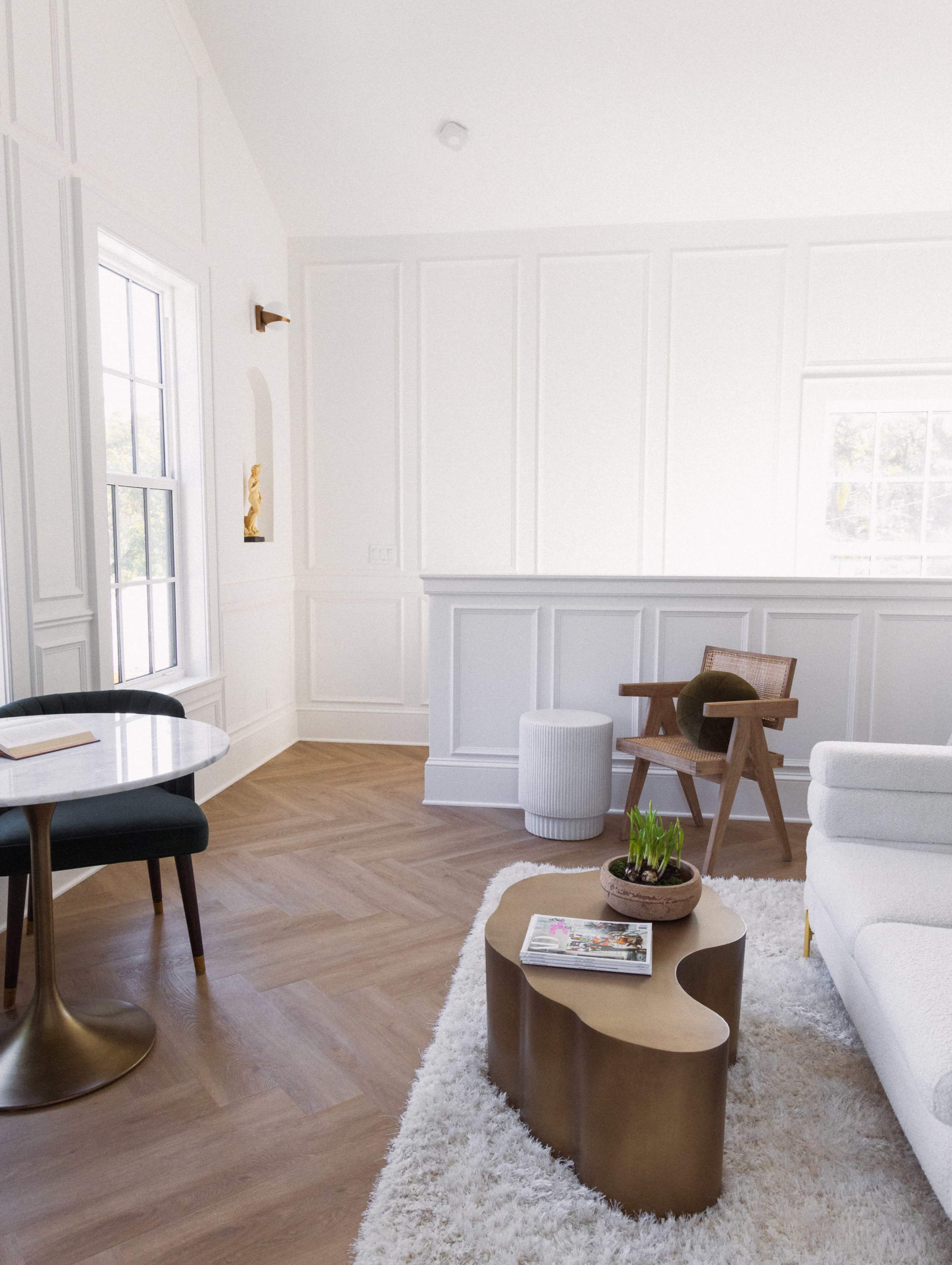 The image depicts a modern living room with light walls, a wooden floor, a round dining table, a decorative coffee table, and a minimalist armchair.