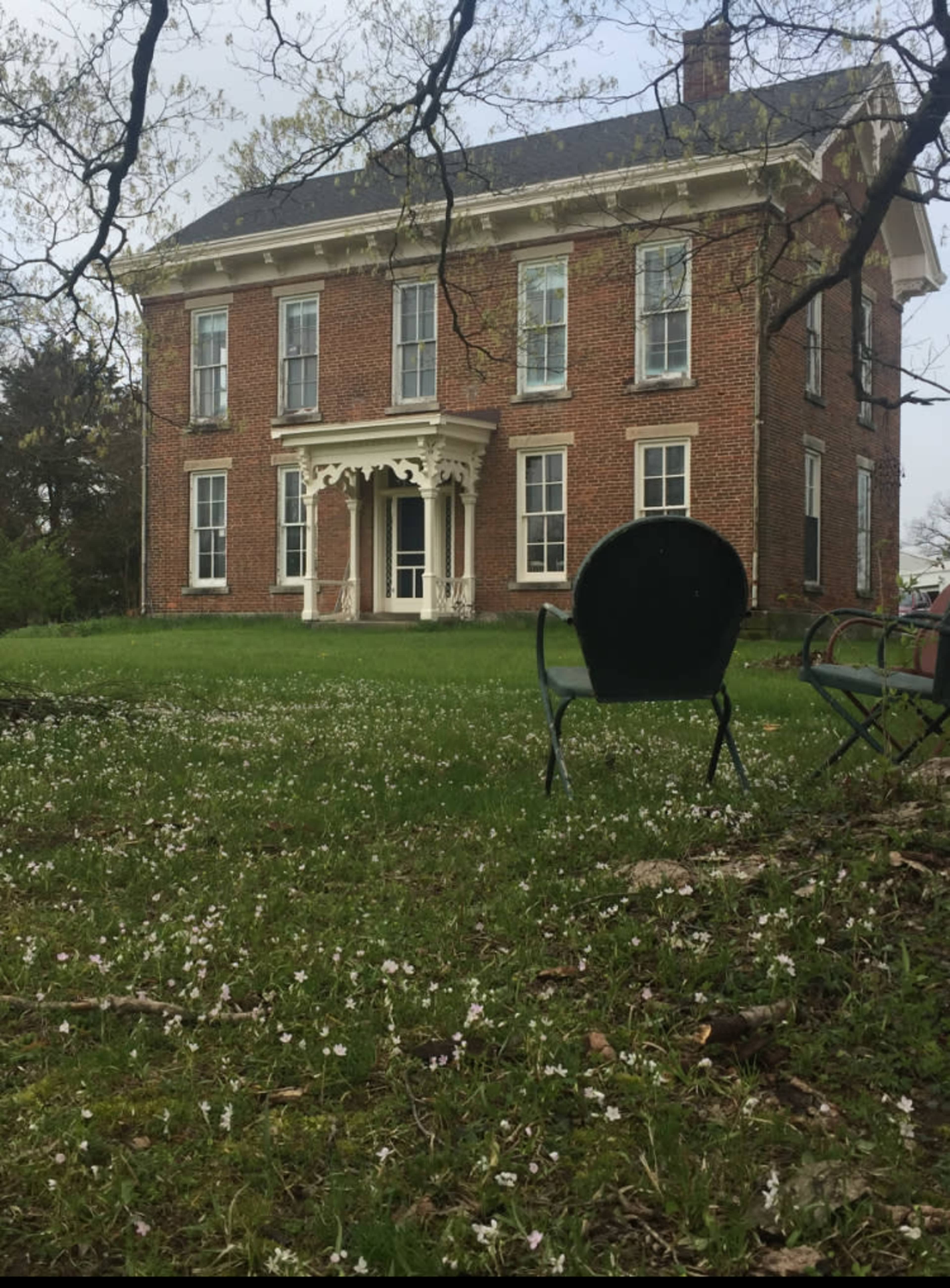 A large brick house with white trim stands in a grassy yard scattered with white flowers, and two green chairs are positioned in the foreground.