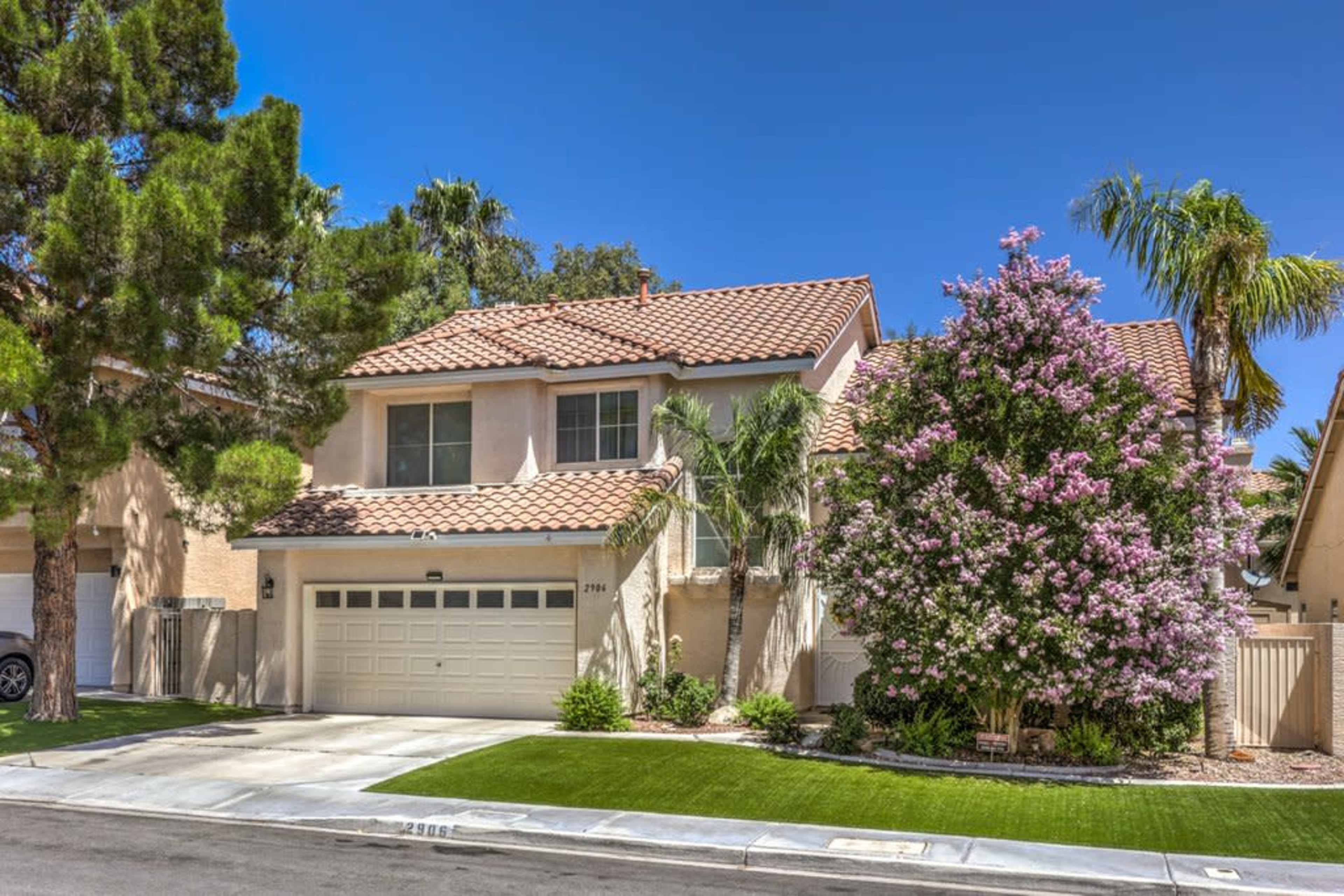 The image shows a two-story house with a tiled roof, surrounded by palm trees and a flowering bush in a residential neighborhood.