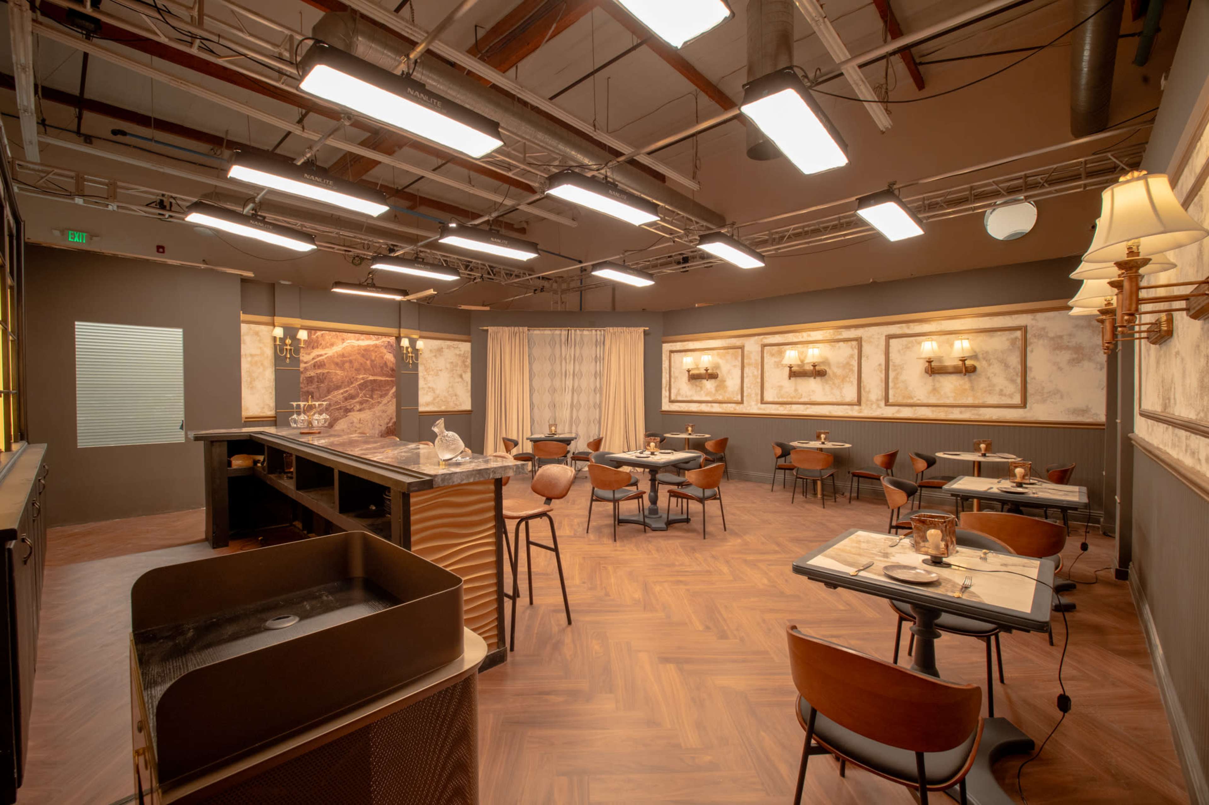 The image shows a spacious restaurant interior with wooden floors, several tables set for dining, and a bar counter illuminated by overhead lights.