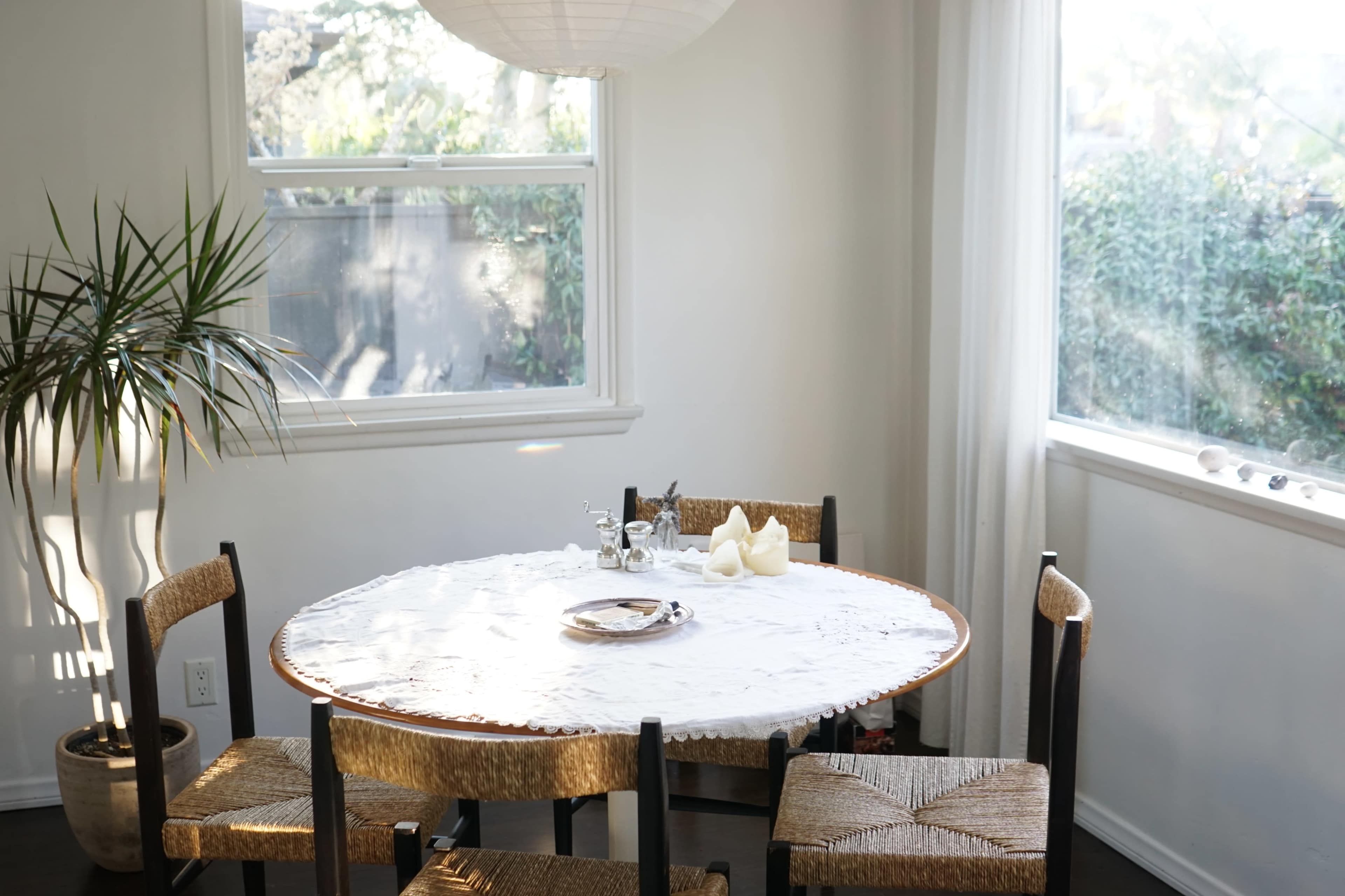A round dining table with a lace tablecloth and simple decor is positioned in a well-lit room with large windows and greenery visible outside.
