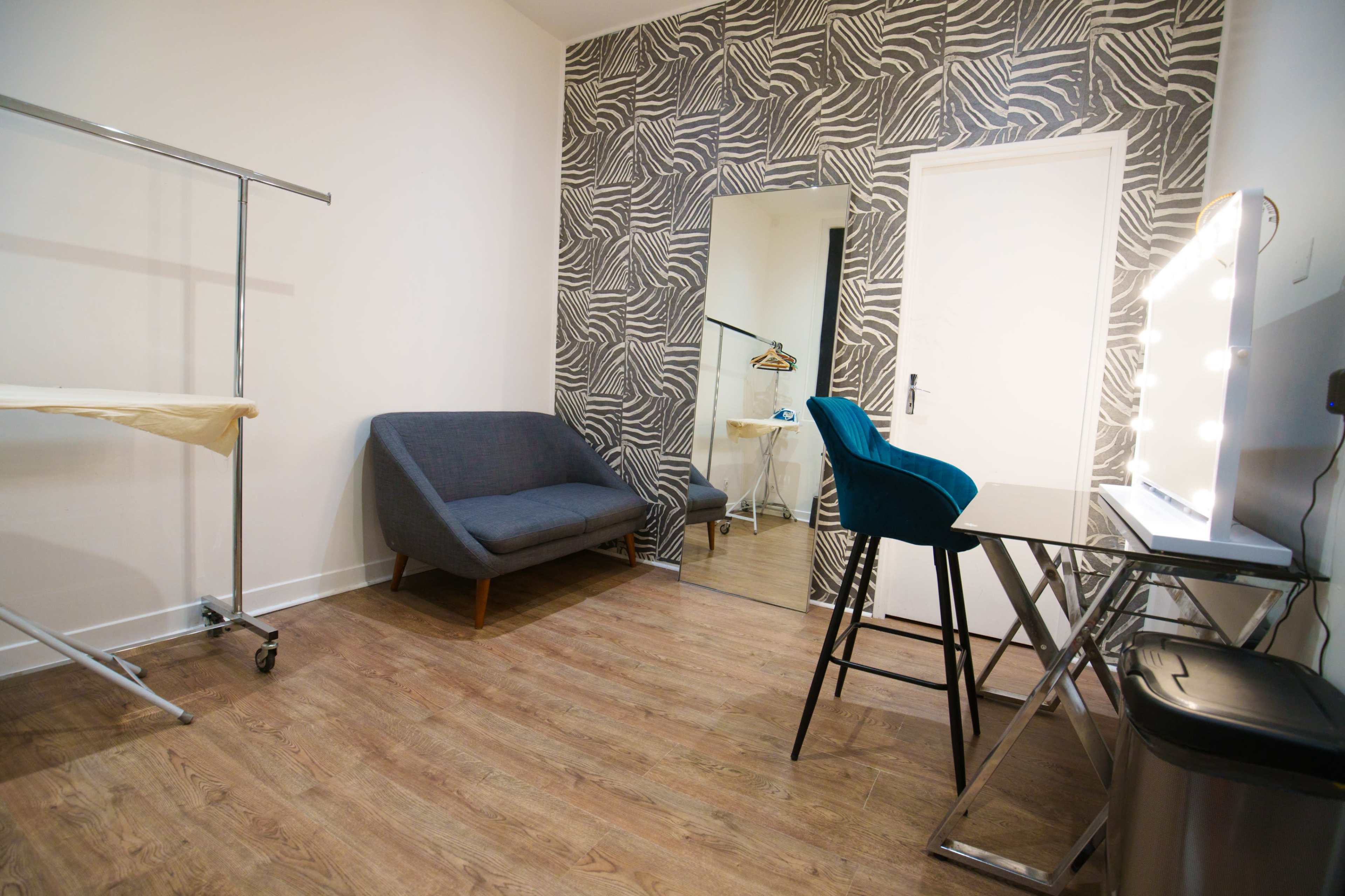 The image shows a small room with a patterned wall, a gray sofa, a mirror, a blue chair, a vanity table, and a clothing rack.