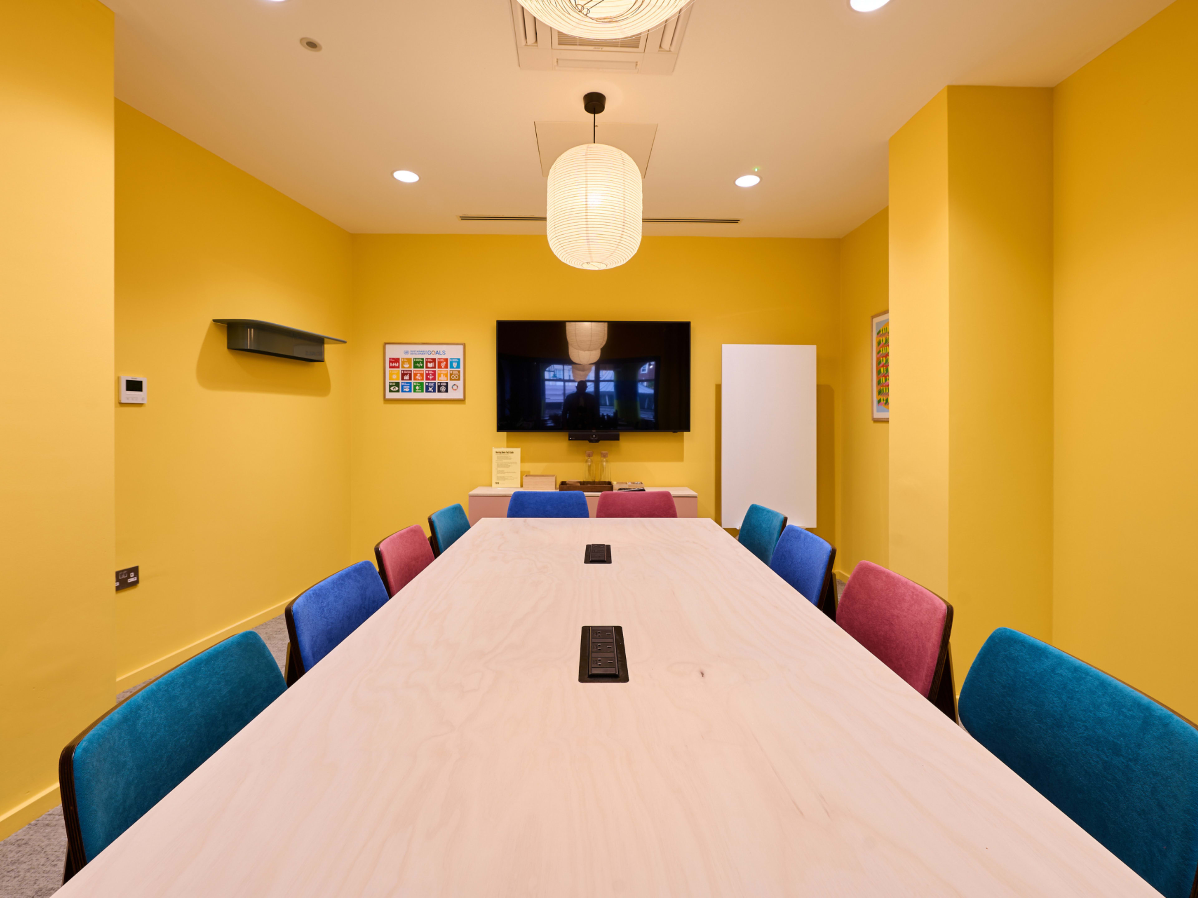 A well-lit meeting room features a long wooden table surrounded by colorful chairs, with a large screen mounted on the yellow wall.