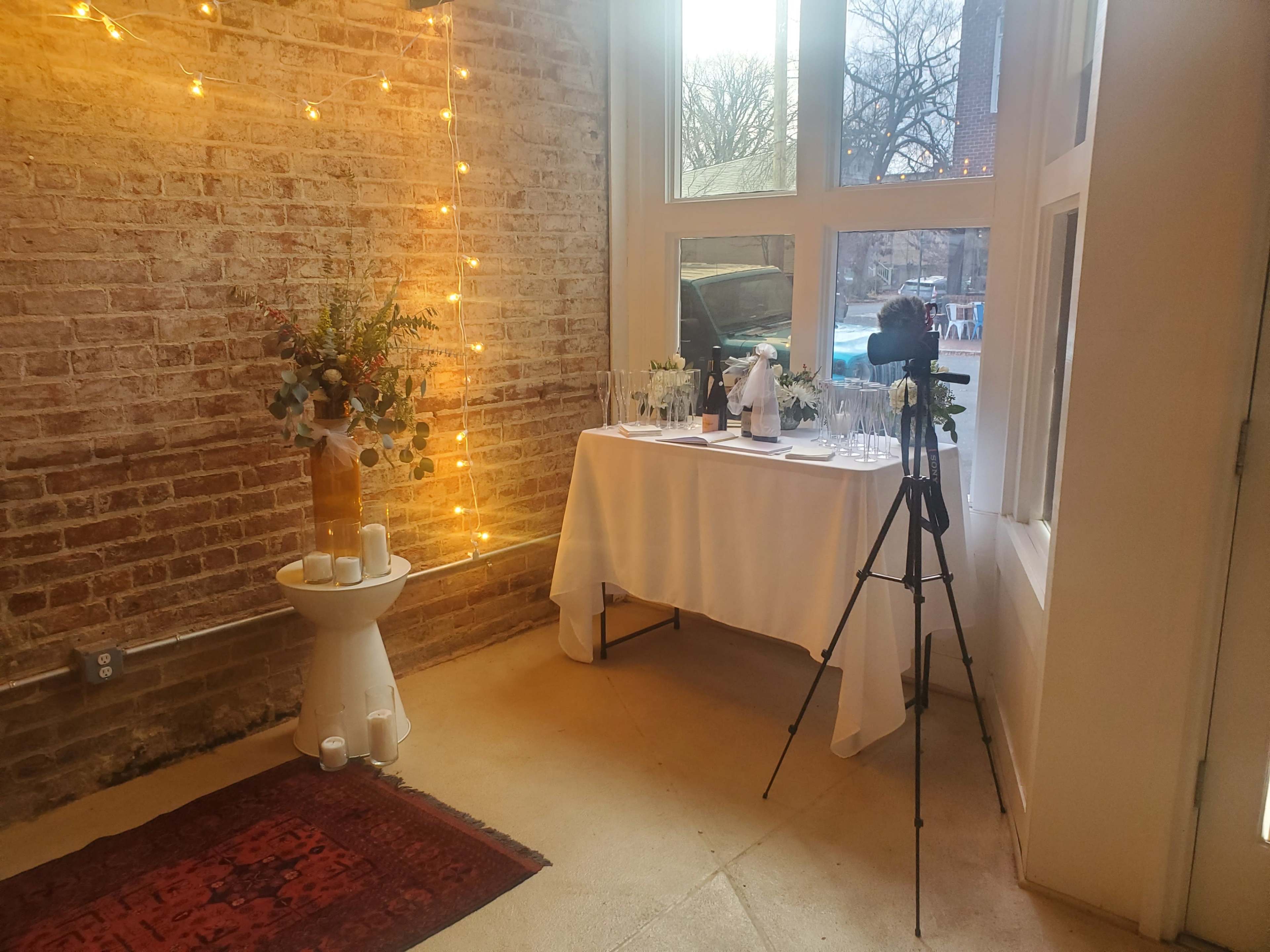The scene features a decorated corner with a table set for an event, adorned with a floral arrangement, candles, and a camera on a tripod beside a window.
