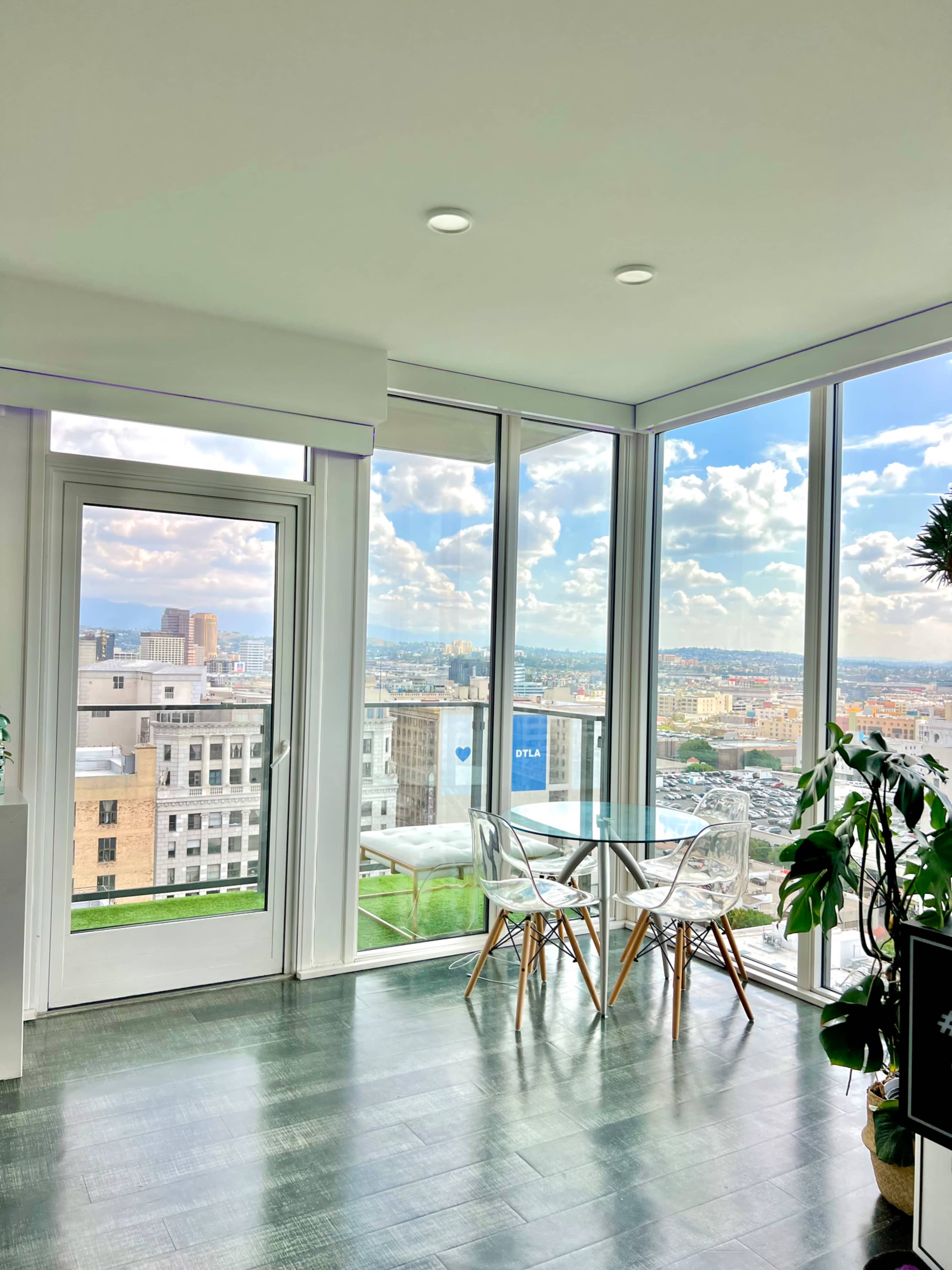 A modern dining area features a clear glass table and chairs alongside large windows overlooking a city skyline and clouds.