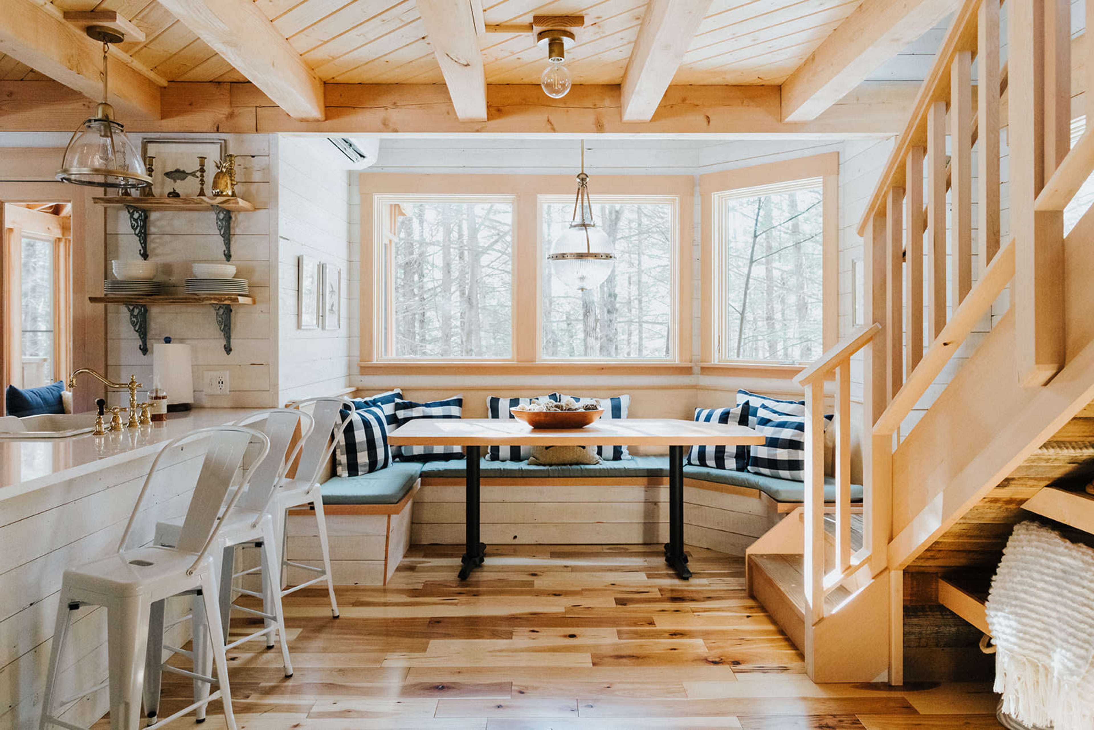 A cozy cabin interior features a dining area with a wooden table surrounded by benches and large windows displaying a forest view.