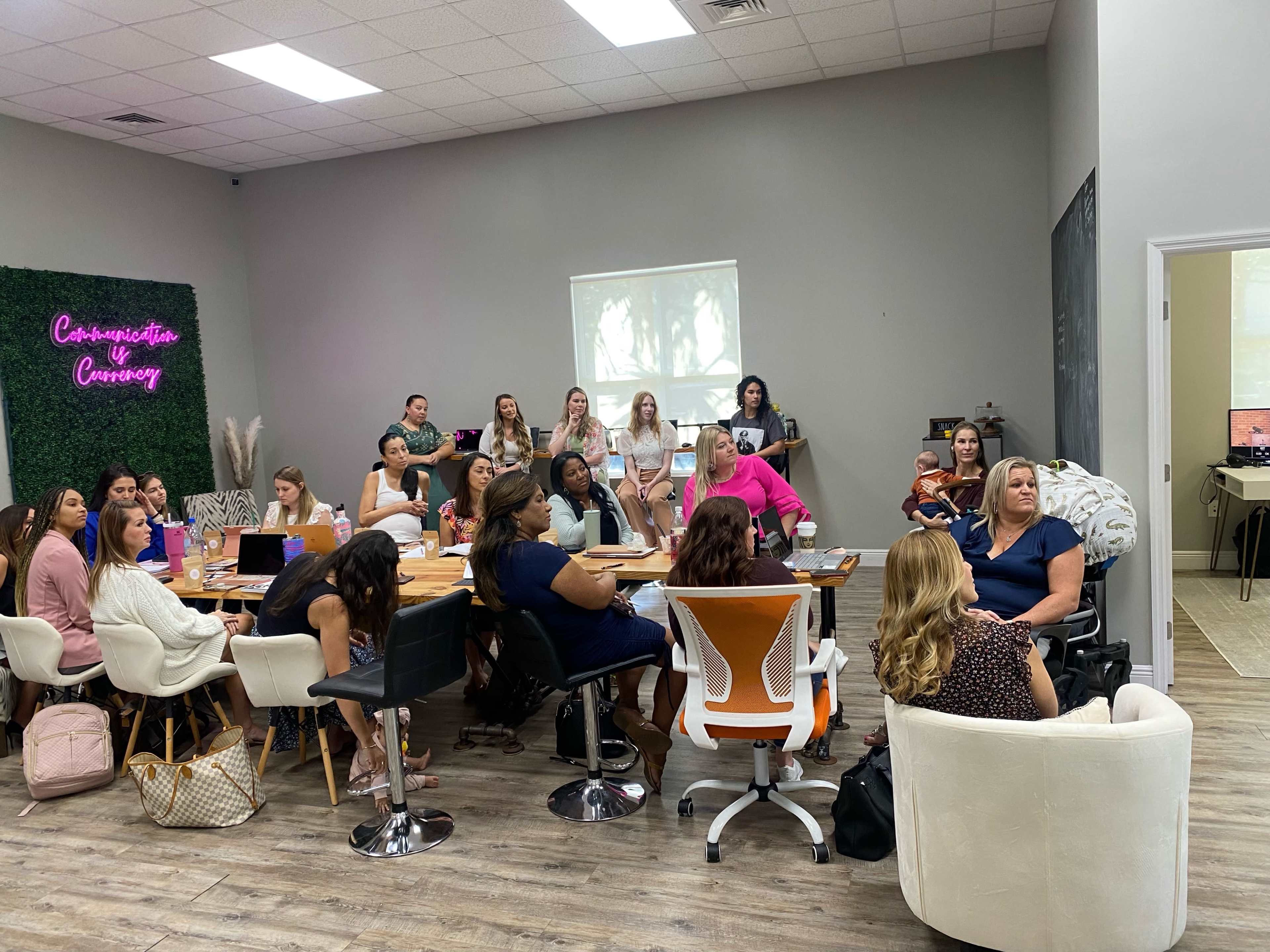 A group of people is gathered around a large table in a bright, modern meeting room, engaging in discussion while some use laptops and a baby is present.