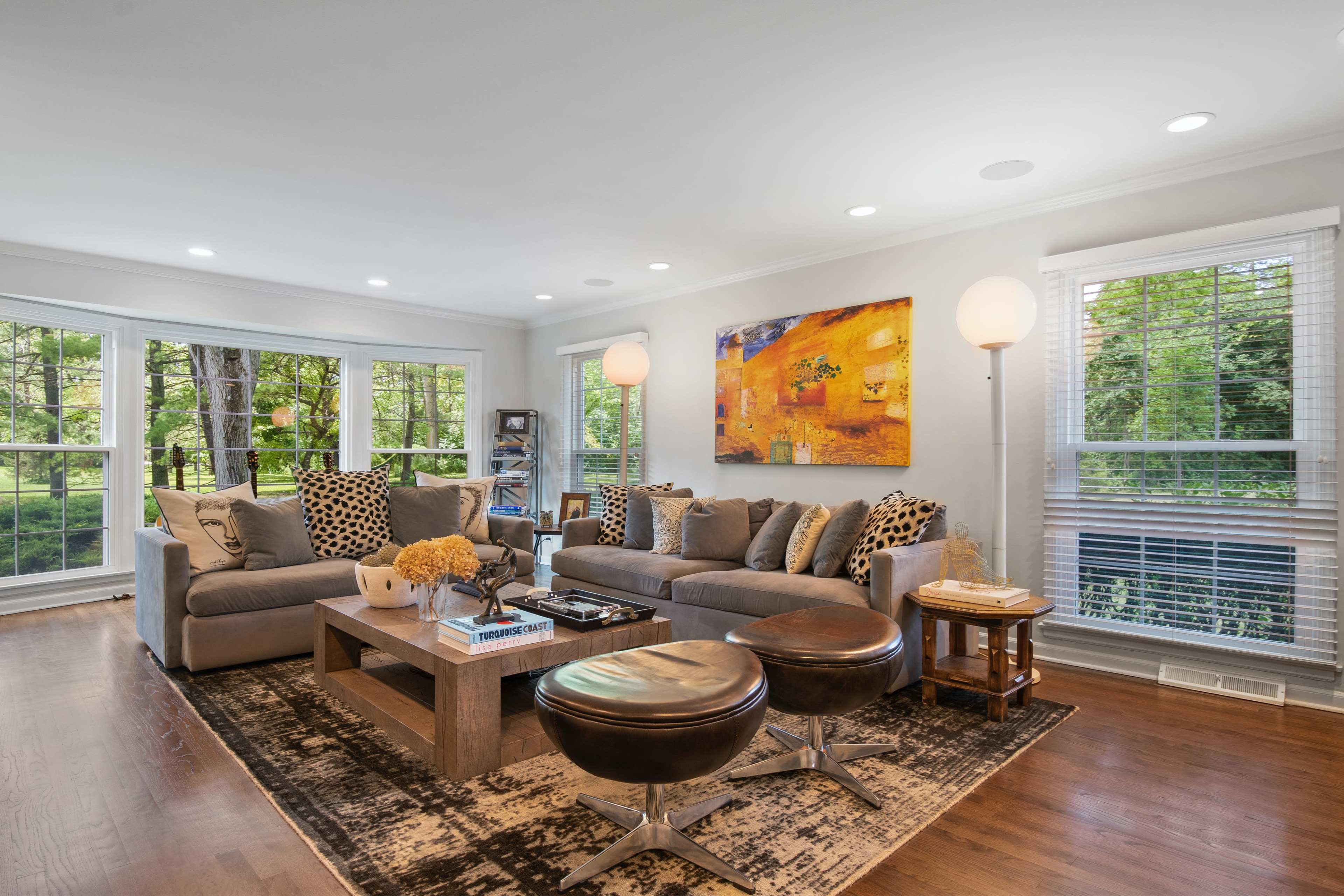 A well-lit living room features two gray sofas, a coffee table, and two dark wooden stools, with large windows offering views of a landscaped yard.
