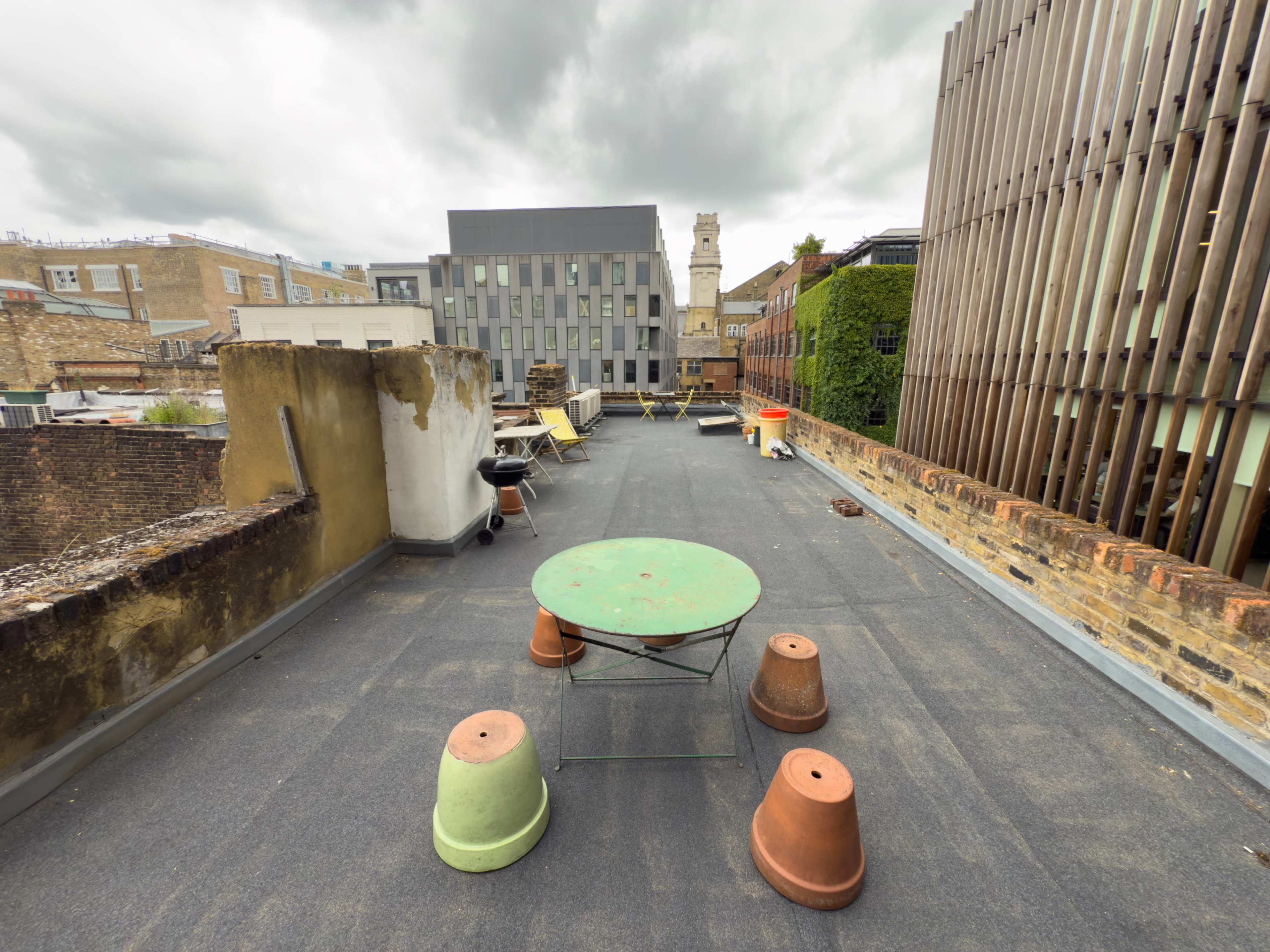 The image shows a rooftop space featuring several colorful tables and planters arranged on a flat surface with buildings in the background.