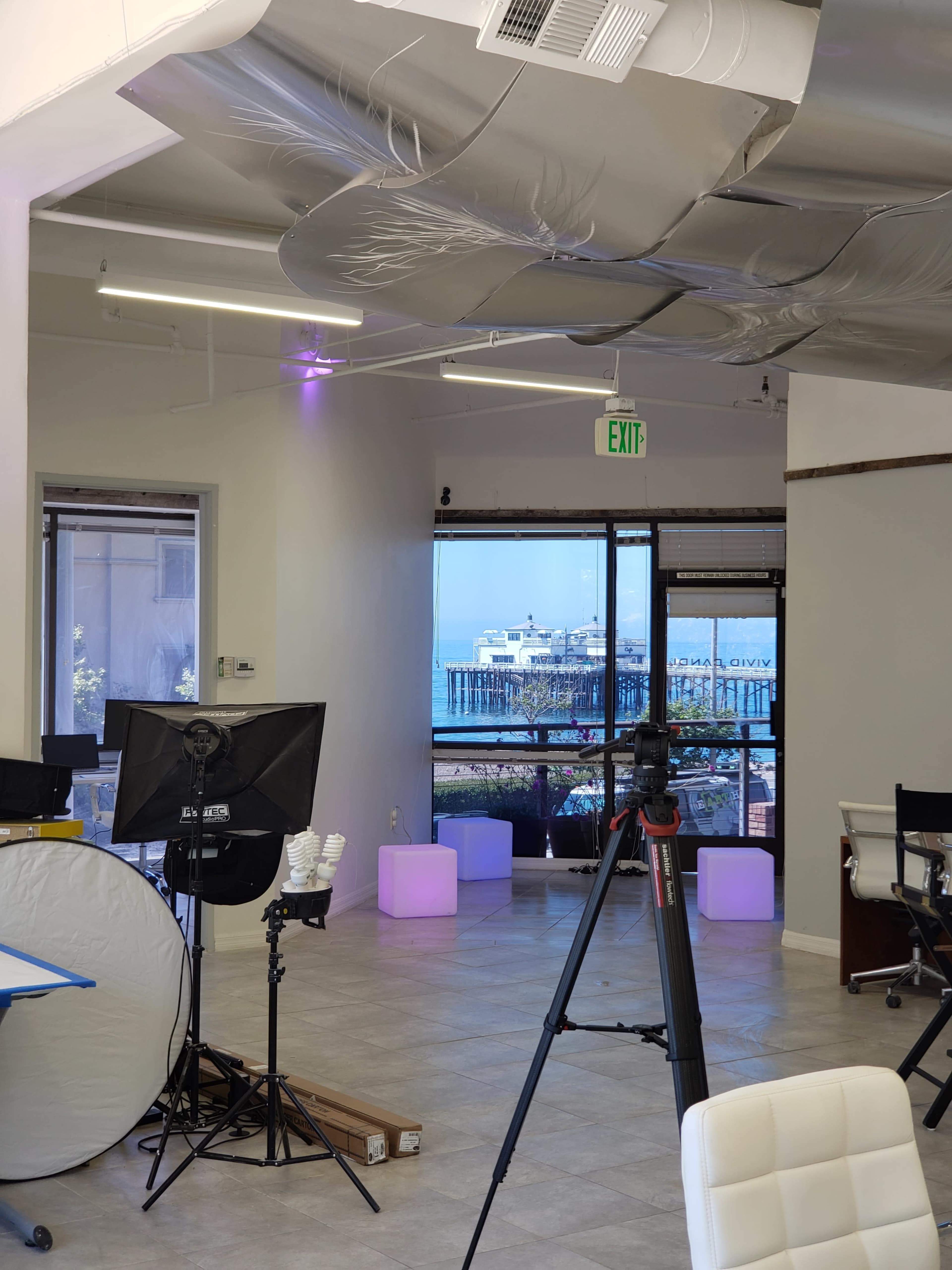 An office space with a view of a pier through large windows, featuring modern lighting and photography equipment set up inside.