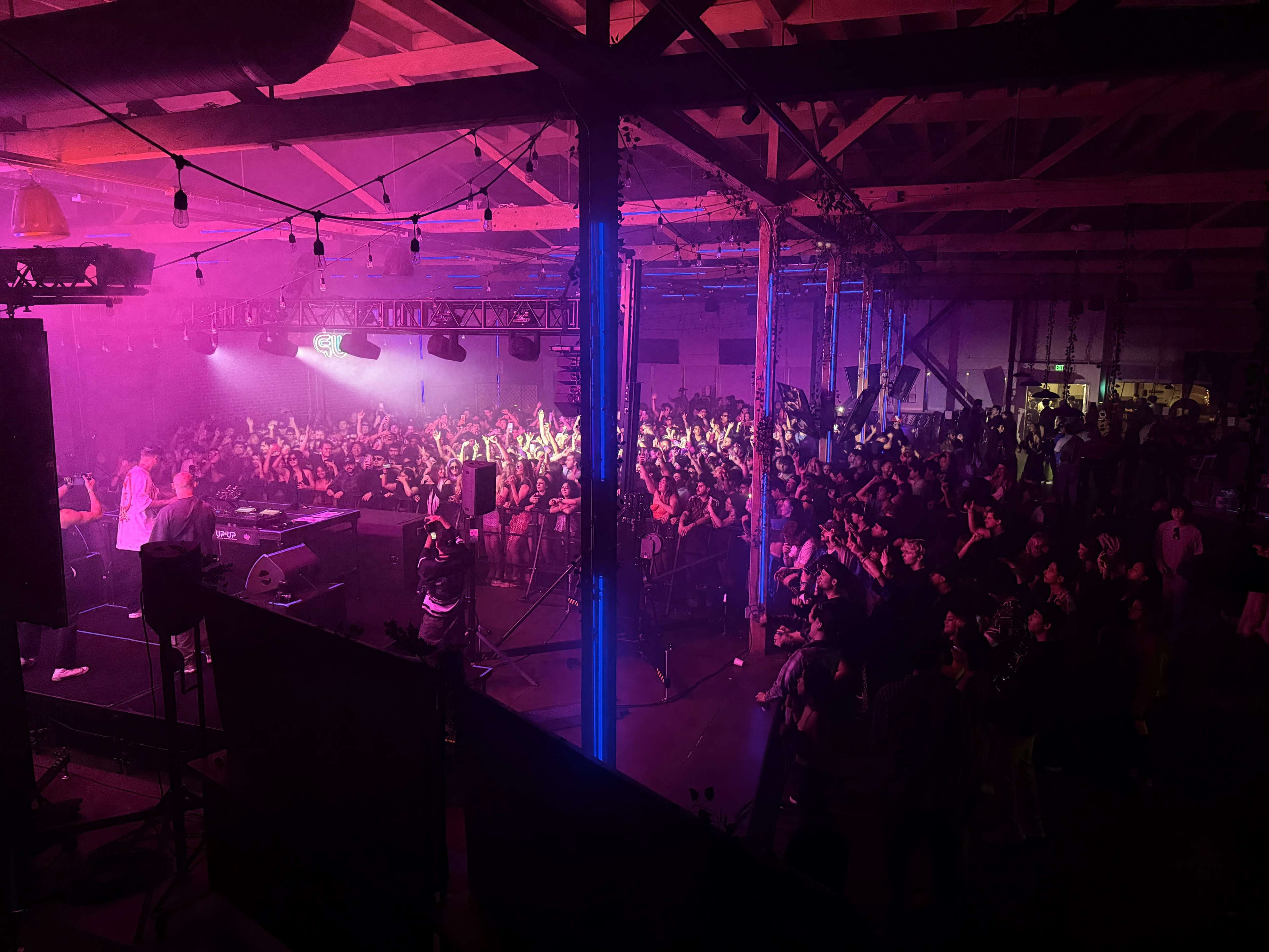 A large crowd is gathered in a dimly lit warehouse for a concert, with colorful lights illuminating the space.