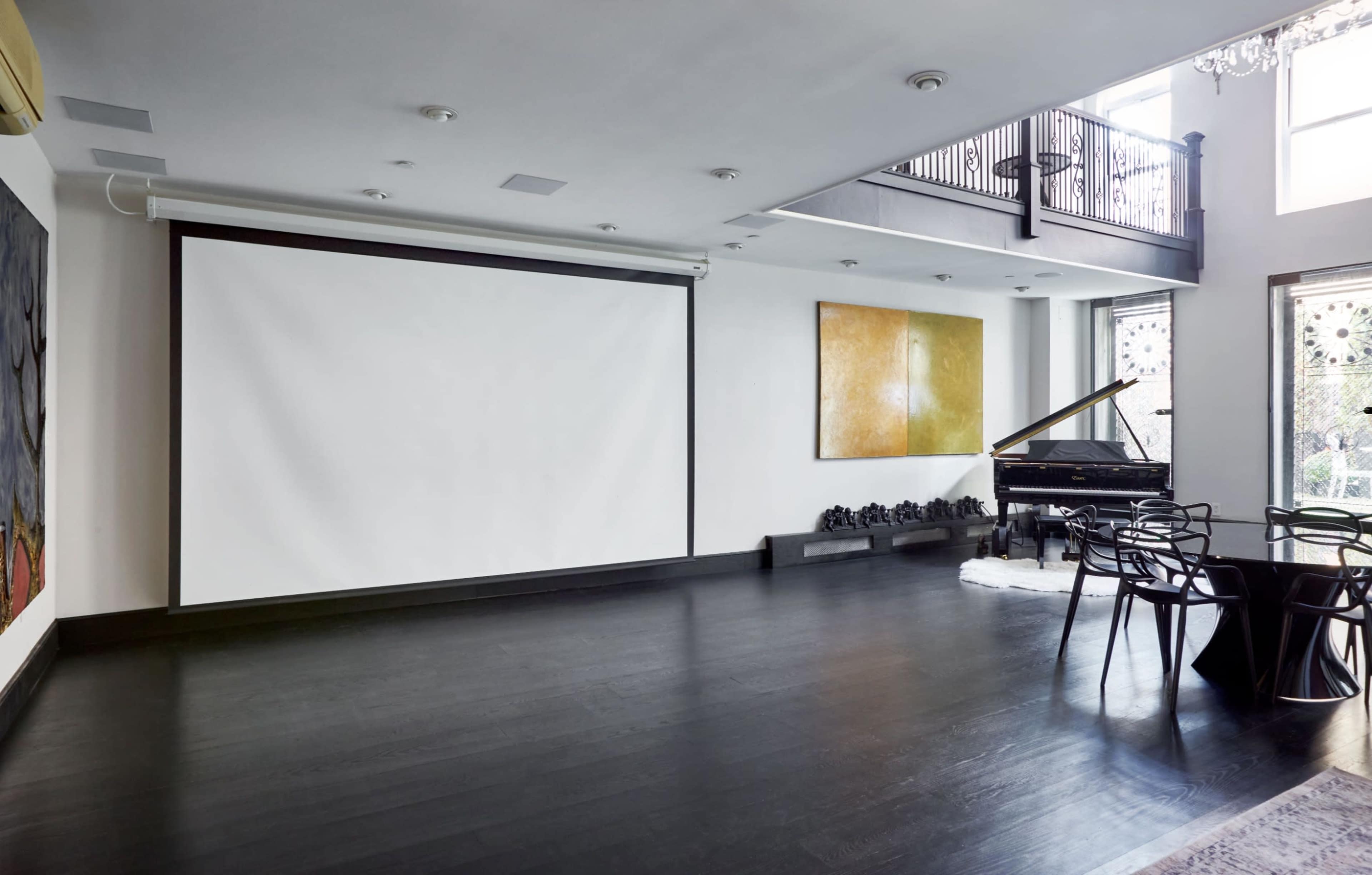 A spacious room with a large blank projector screen, a grand piano, and a circular dining table with black chairs.