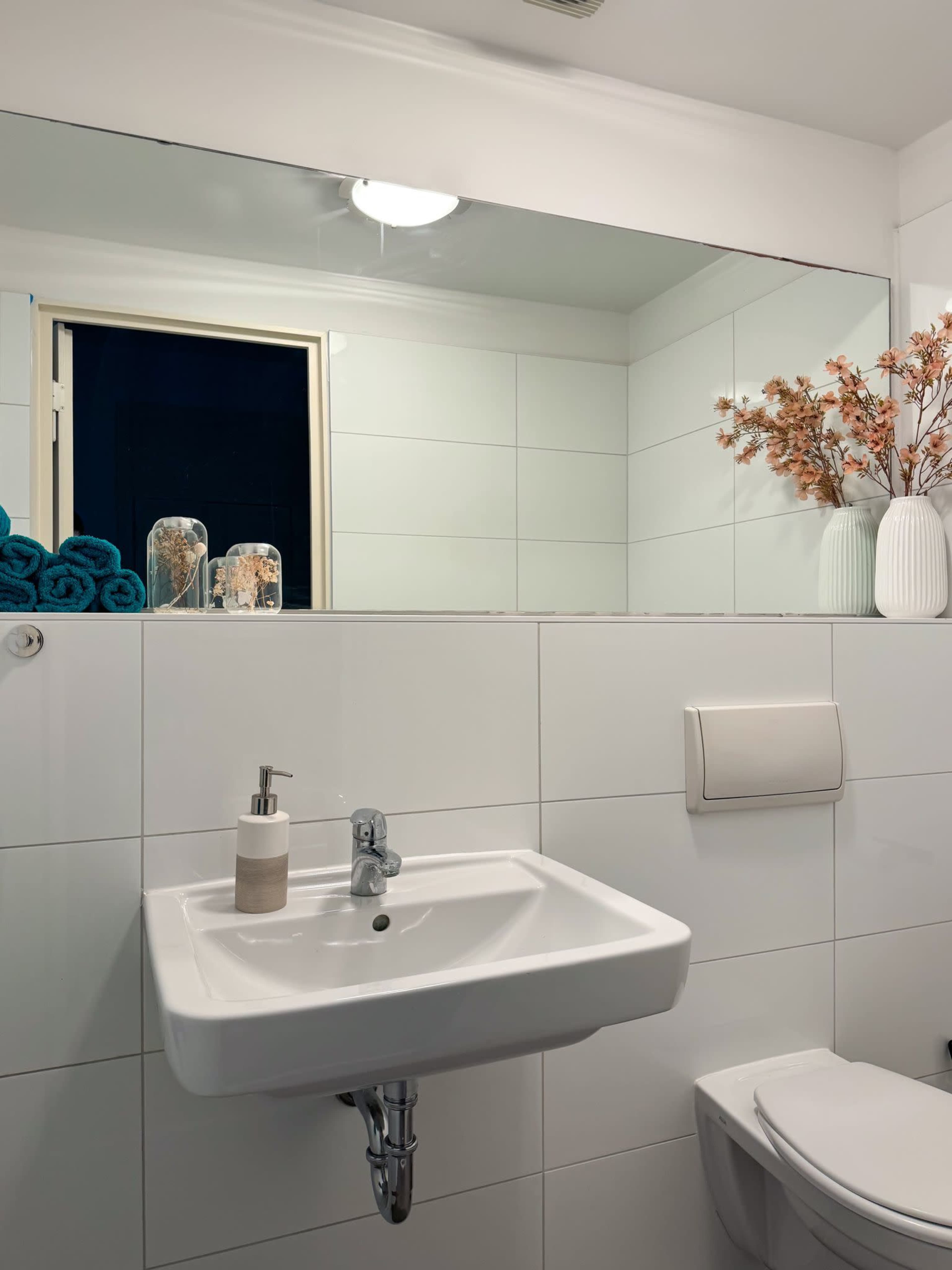 A clean and modern bathroom features a white sink, decorative towels, and a small arrangement of dried flowers on the counter.