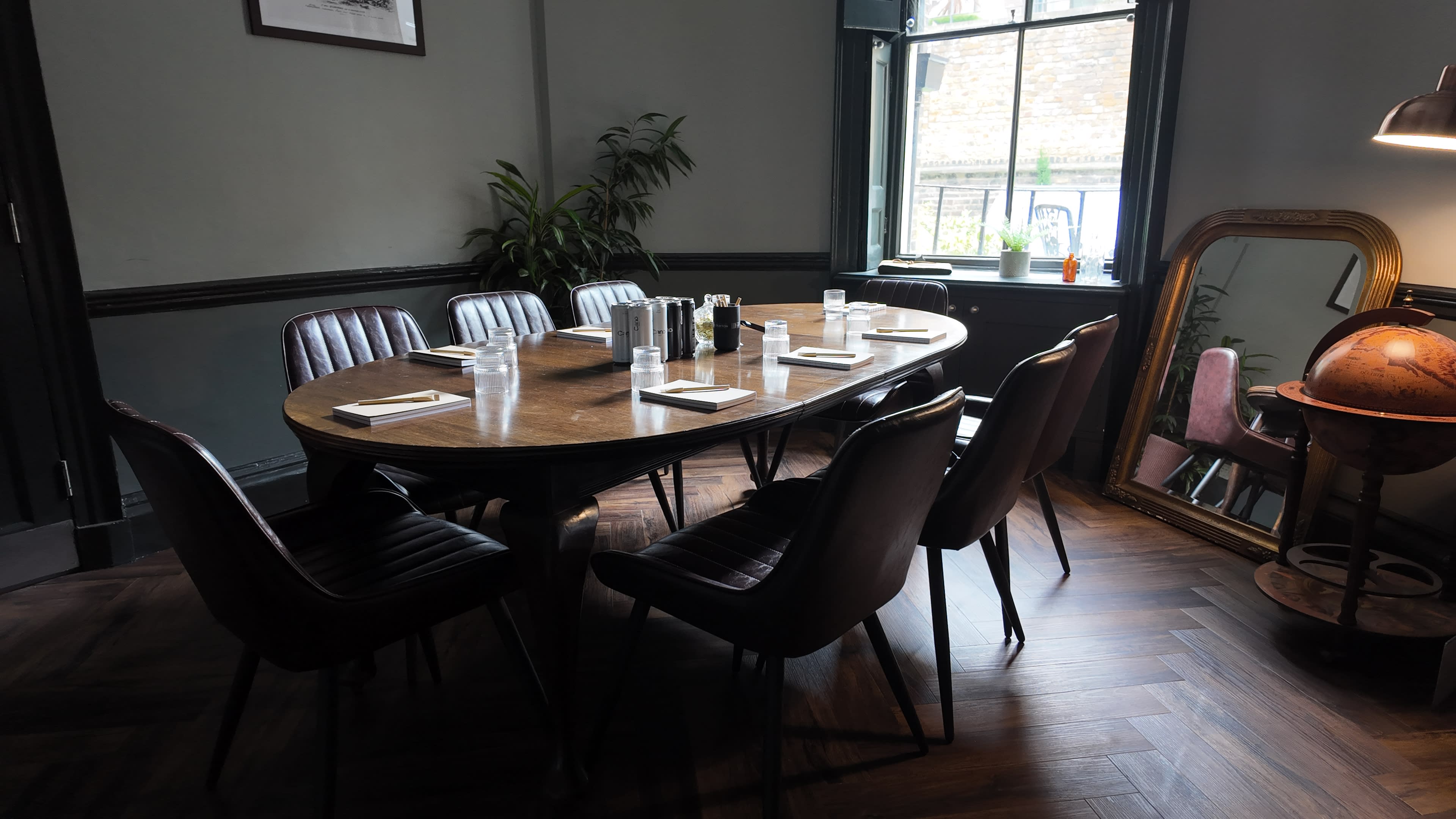 A large wooden table surrounded by black and brown chairs is set up for a meeting in a well-lit room with a window and decorative plants.