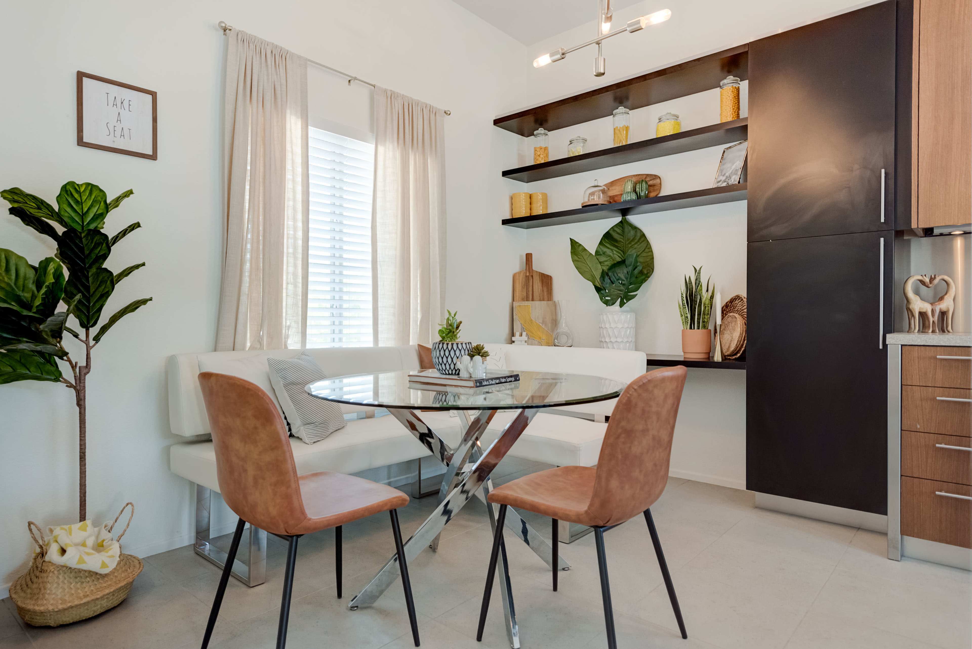 A dining area features a round glass table with a chrome base, surrounded by two brown chairs, near a shelf displaying decor and plants.