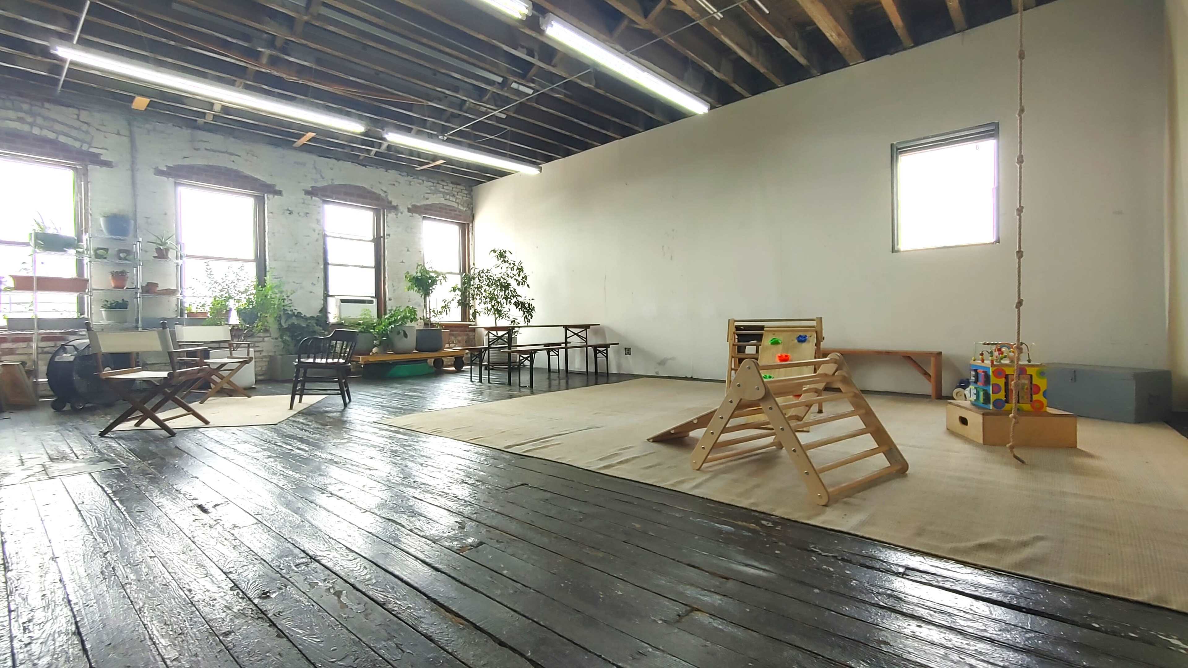 The image shows a spacious room with wooden floors, large windows, and minimal furnishings, including a climbing structure and scattered toys.