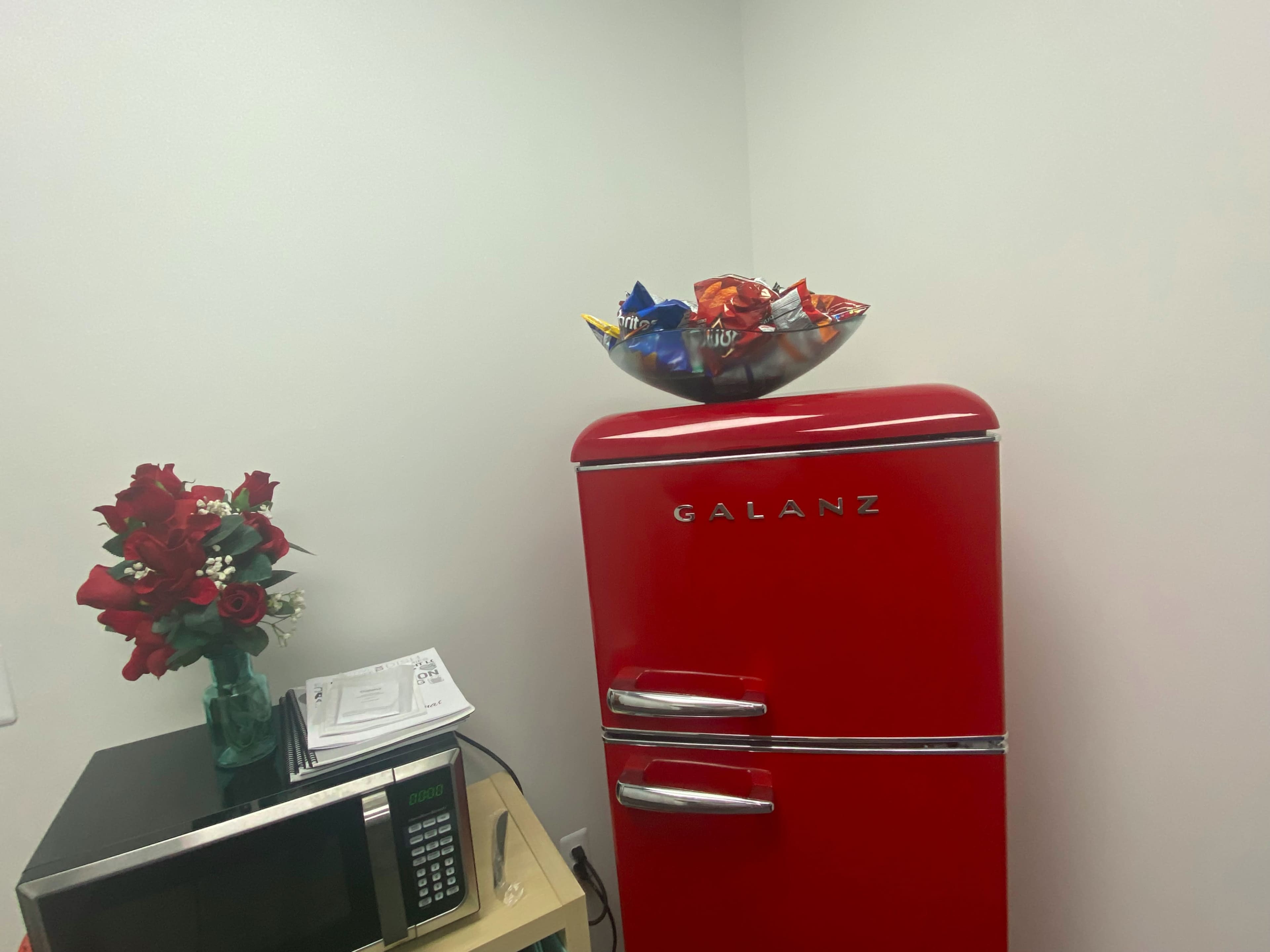 A red Galanz refrigerator stands next to a microwave, with a bowl of snacks on top and a bouquet of roses nearby.