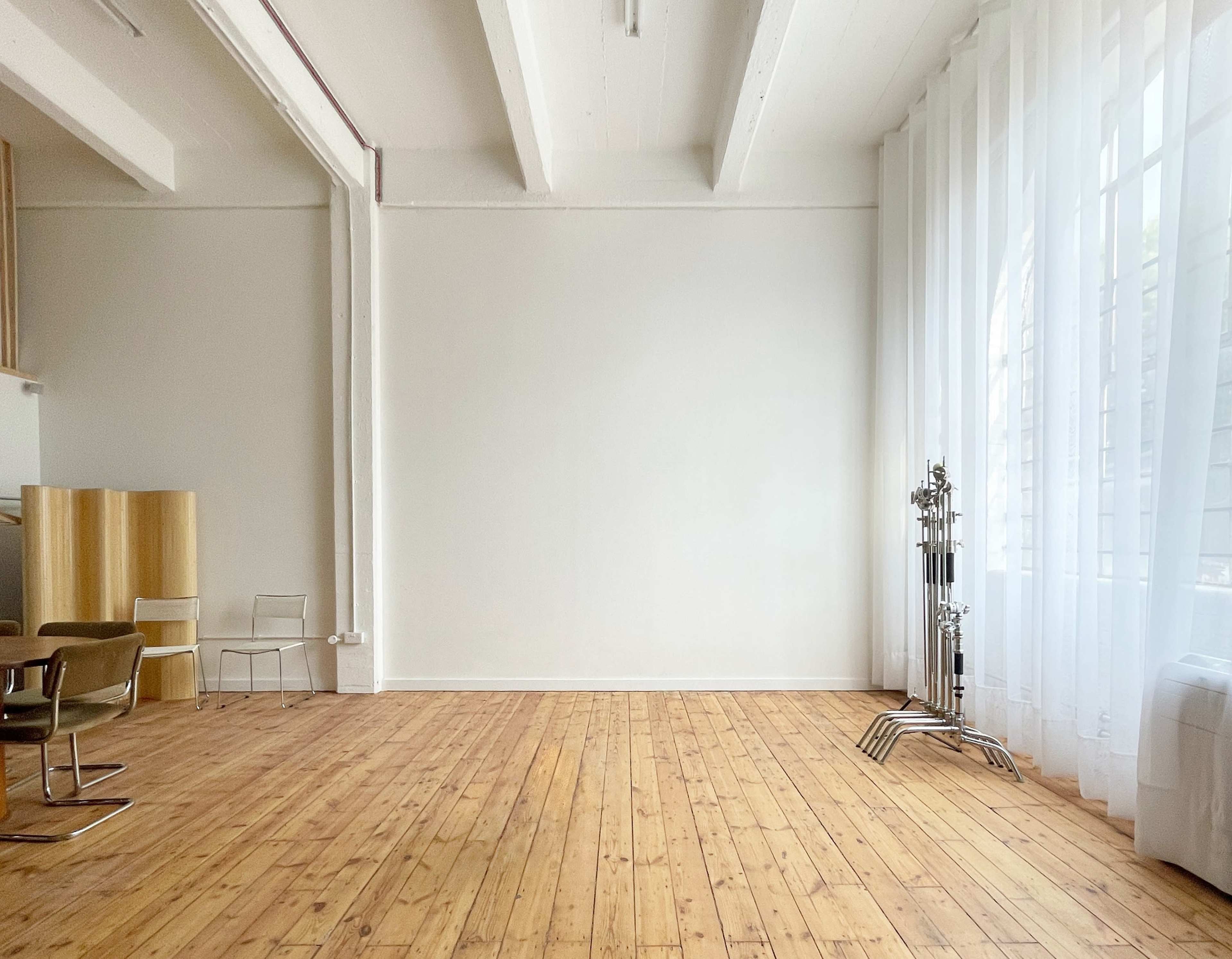 A spacious, minimalist interior with wooden flooring, large windows covered by sheer curtains, and a few pieces of furniture including chairs and a decorative stand.