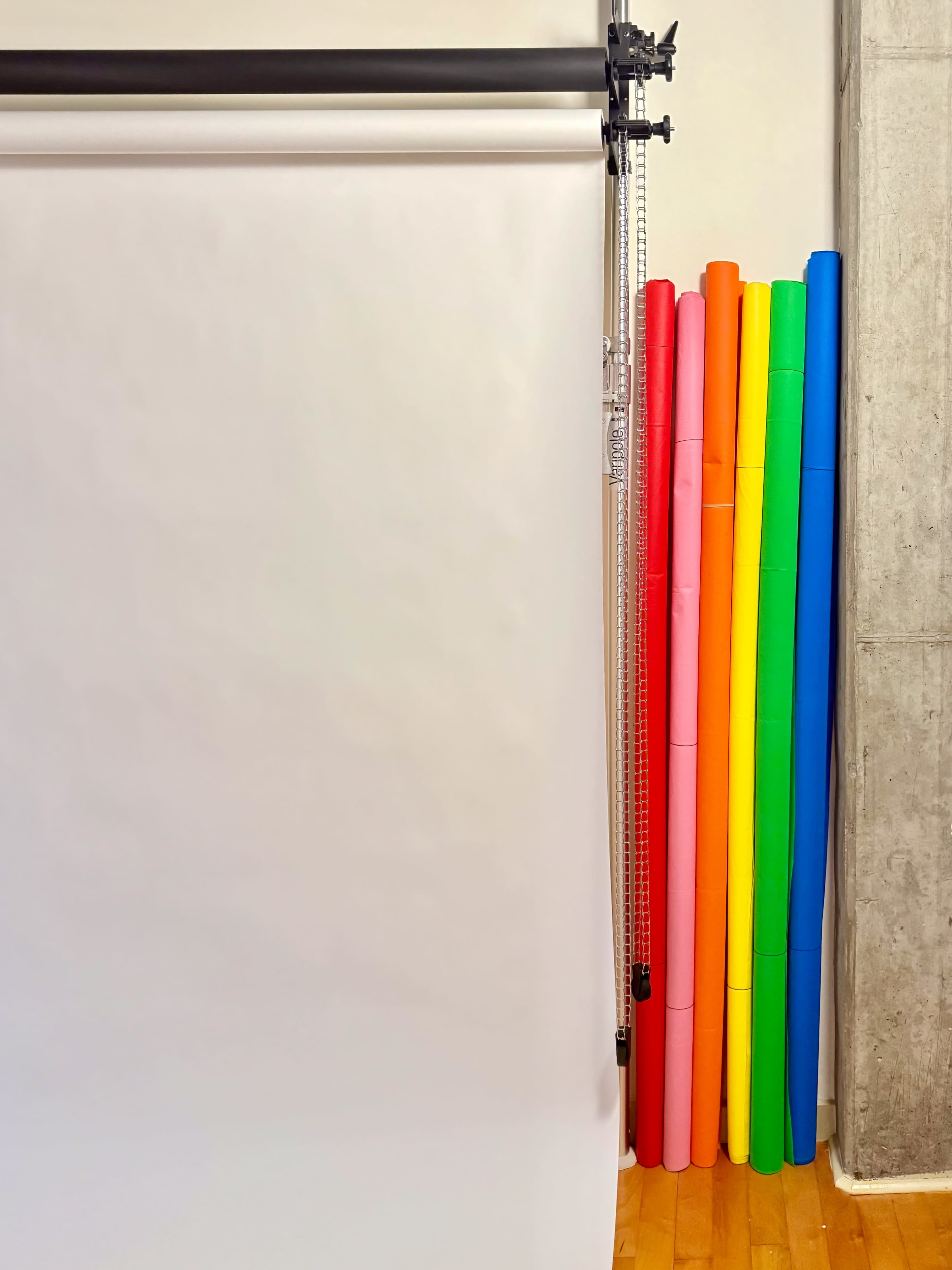 The image shows a bright assortment of rolled backdrop papers in various colors, leaning against a wall next to a white backdrop setup.