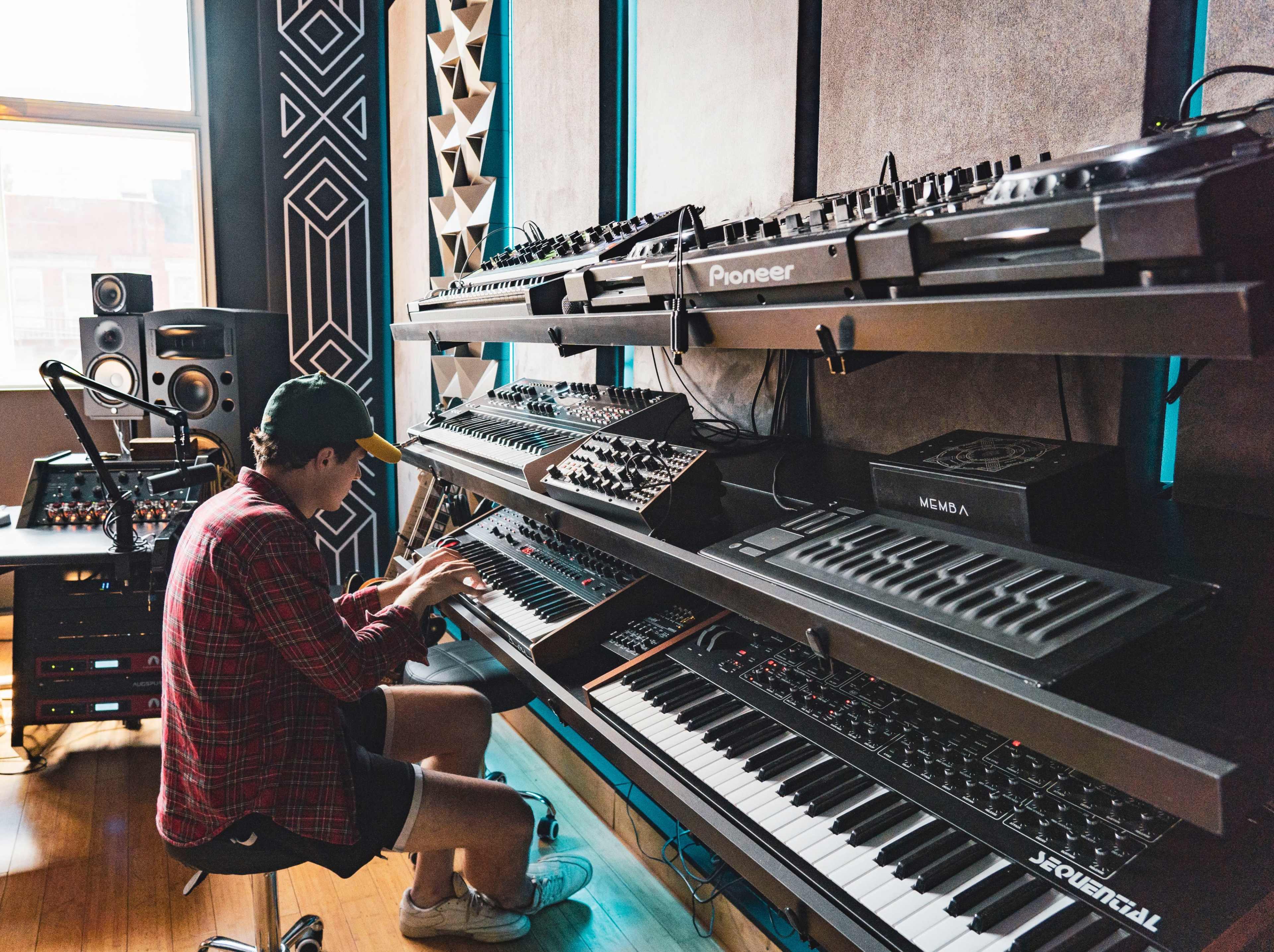 A person wearing a cap is seated on the floor, interacting with a variety of synthesizers and keyboards arranged on metal shelves in a music studio.