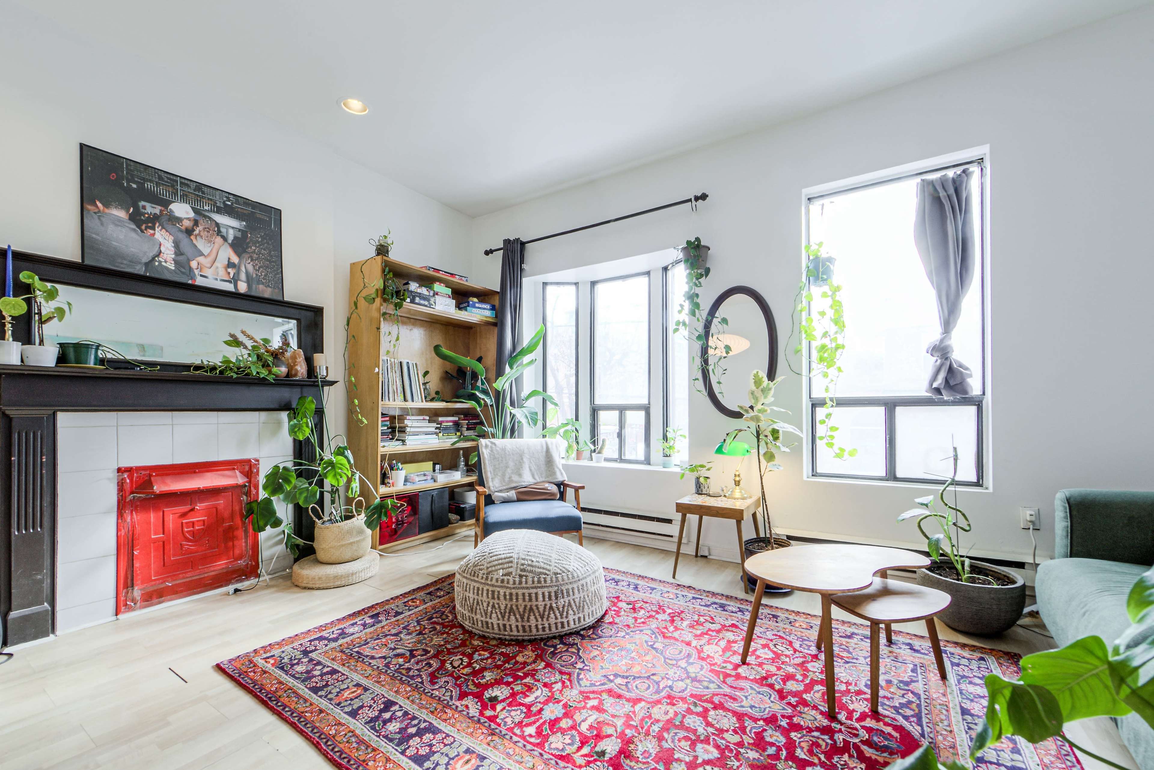 The image shows a bright living room with a red fireplace, a bookshelf filled with plants and books, and a patterned rug under a coffee table.