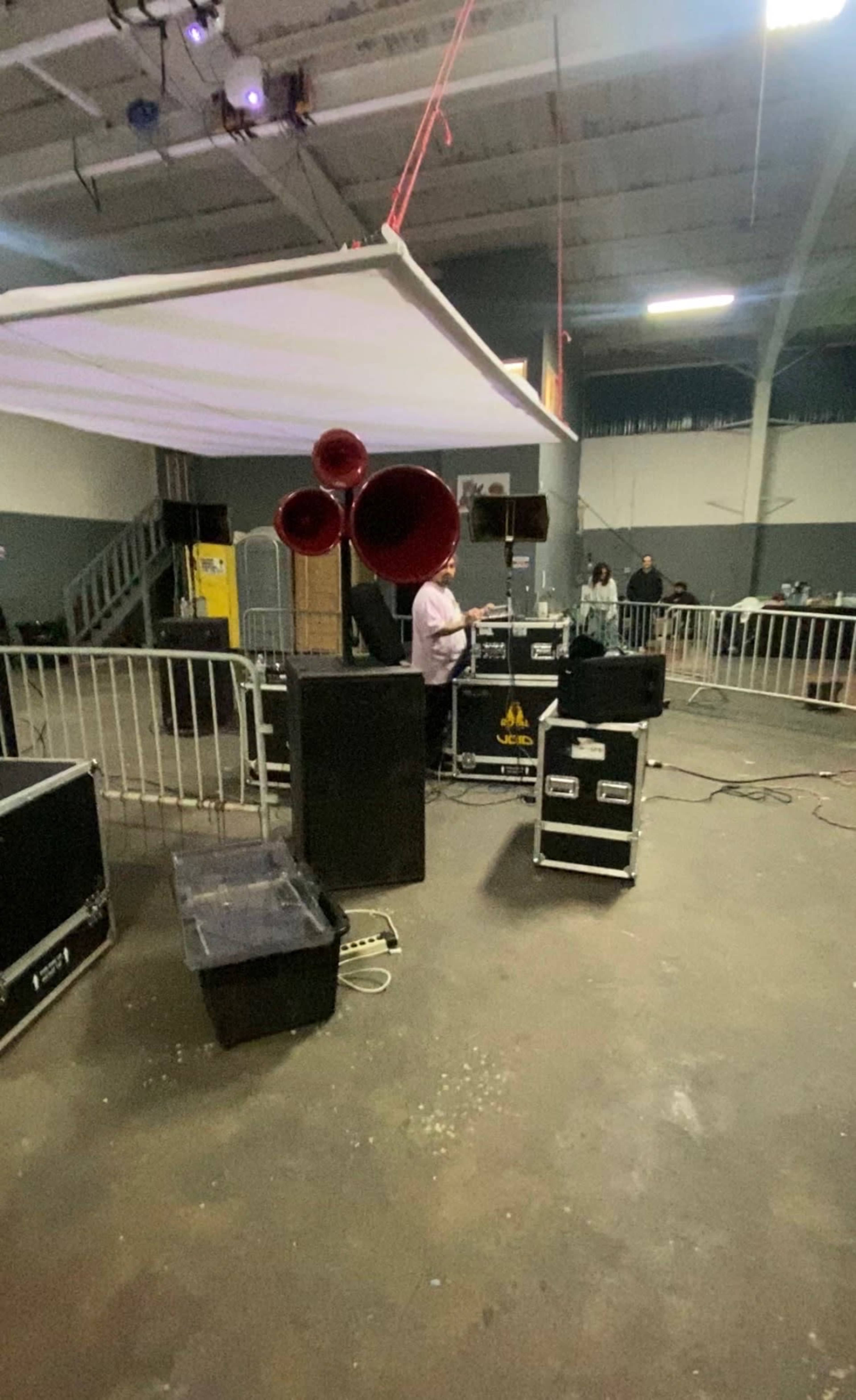 A speaker setup with three large horns is positioned next to a DJ, surrounded by sound equipment and barriers in an indoor venue.