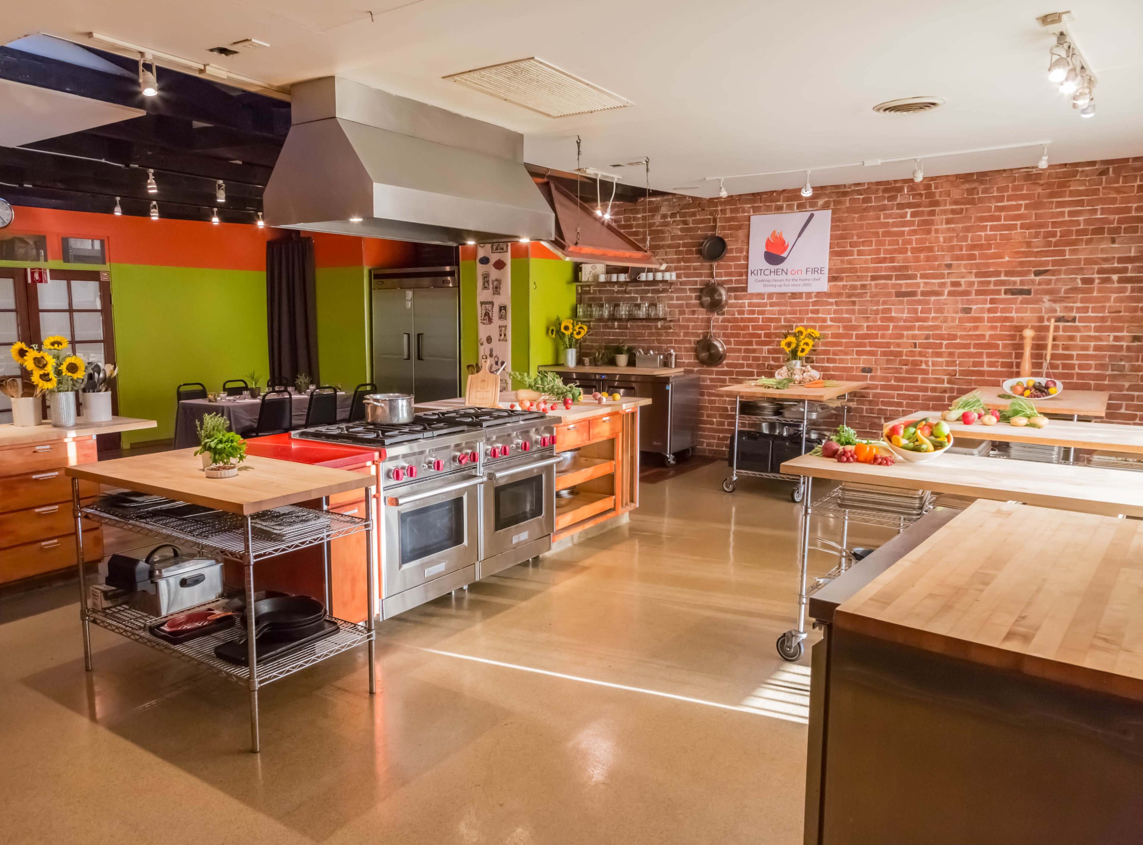 A modern kitchen space with stainless steel appliances, wooden preparation tables, and bright green and brick walls, featuring sunflowers as a decorative element.