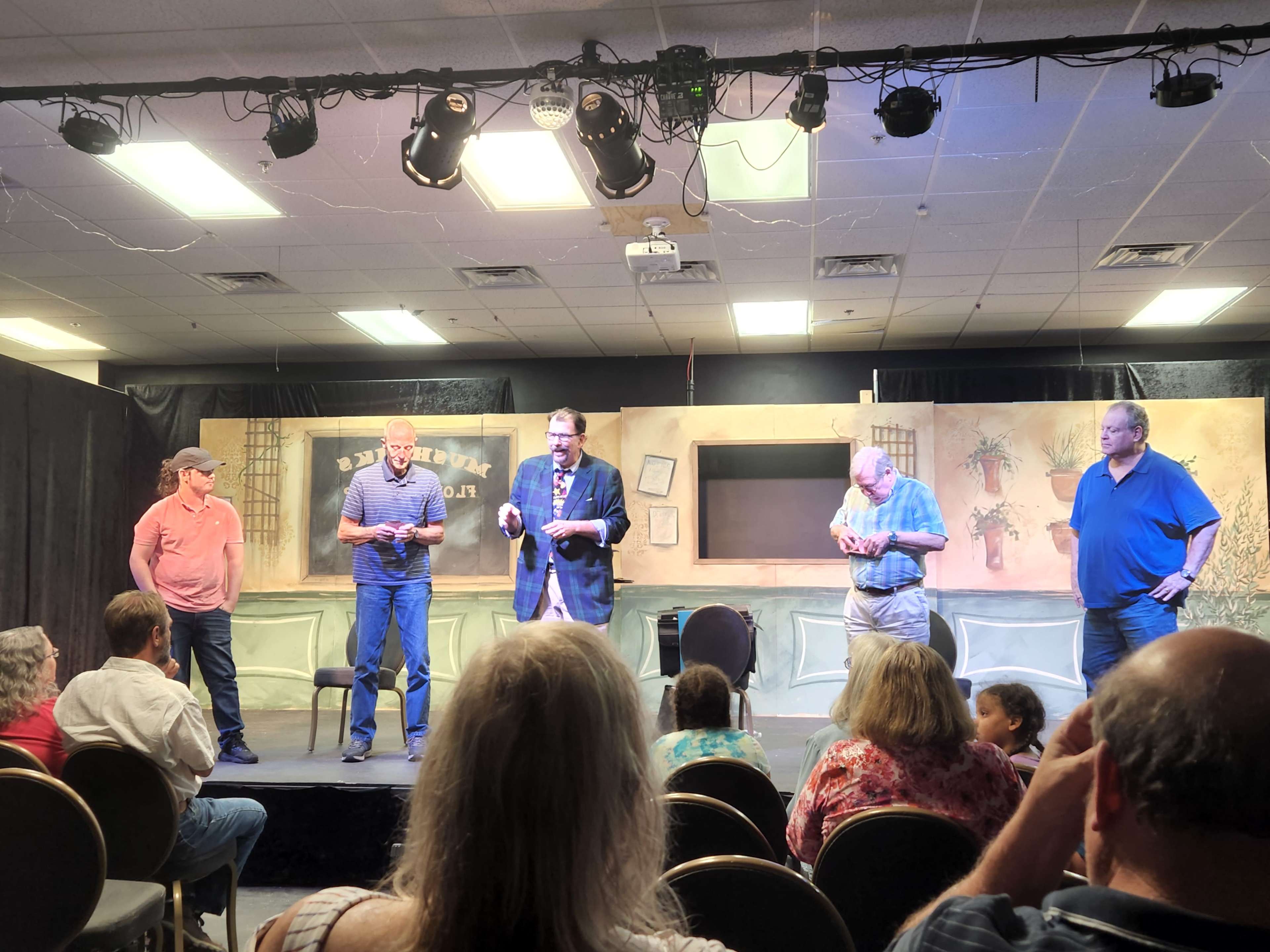 A group of five men are standing on a stage, engaging with each other while an audience observes from their seats.