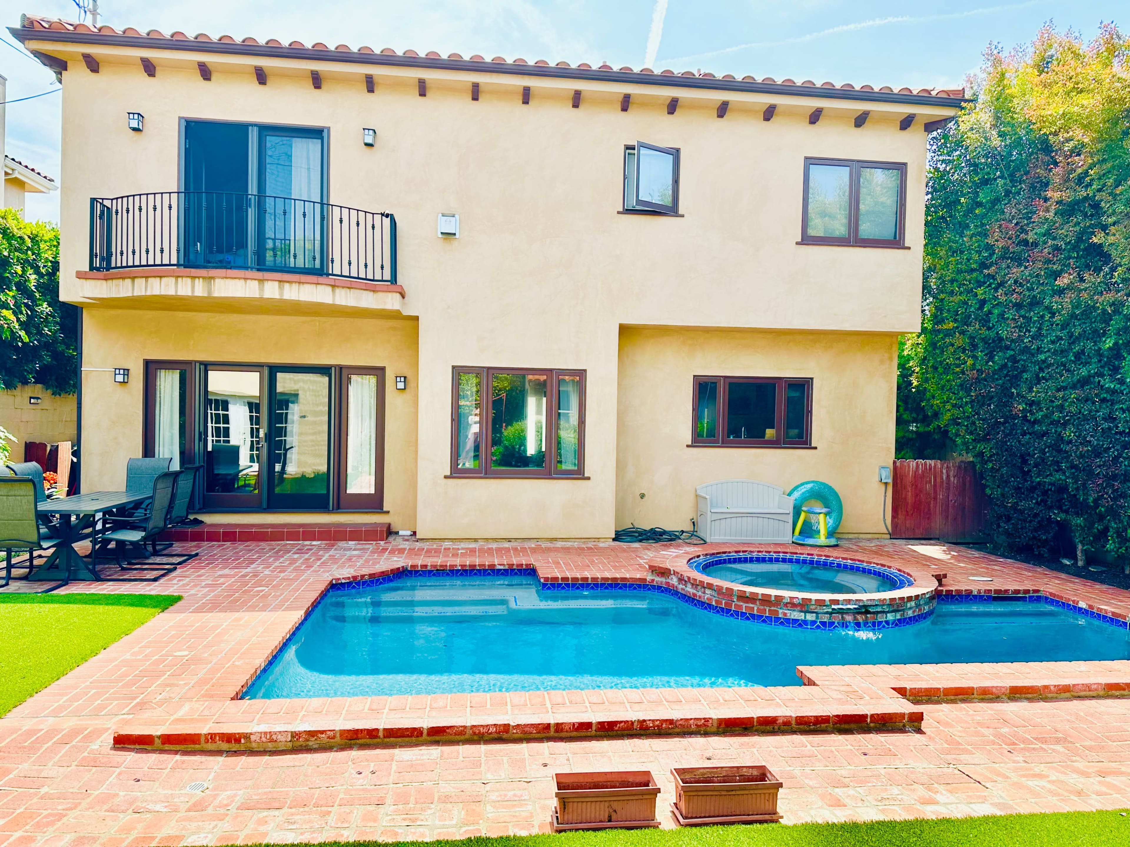 A two-story house with a patio area, a blue swimming pool, and a spa surrounded by brick paving and greenery.