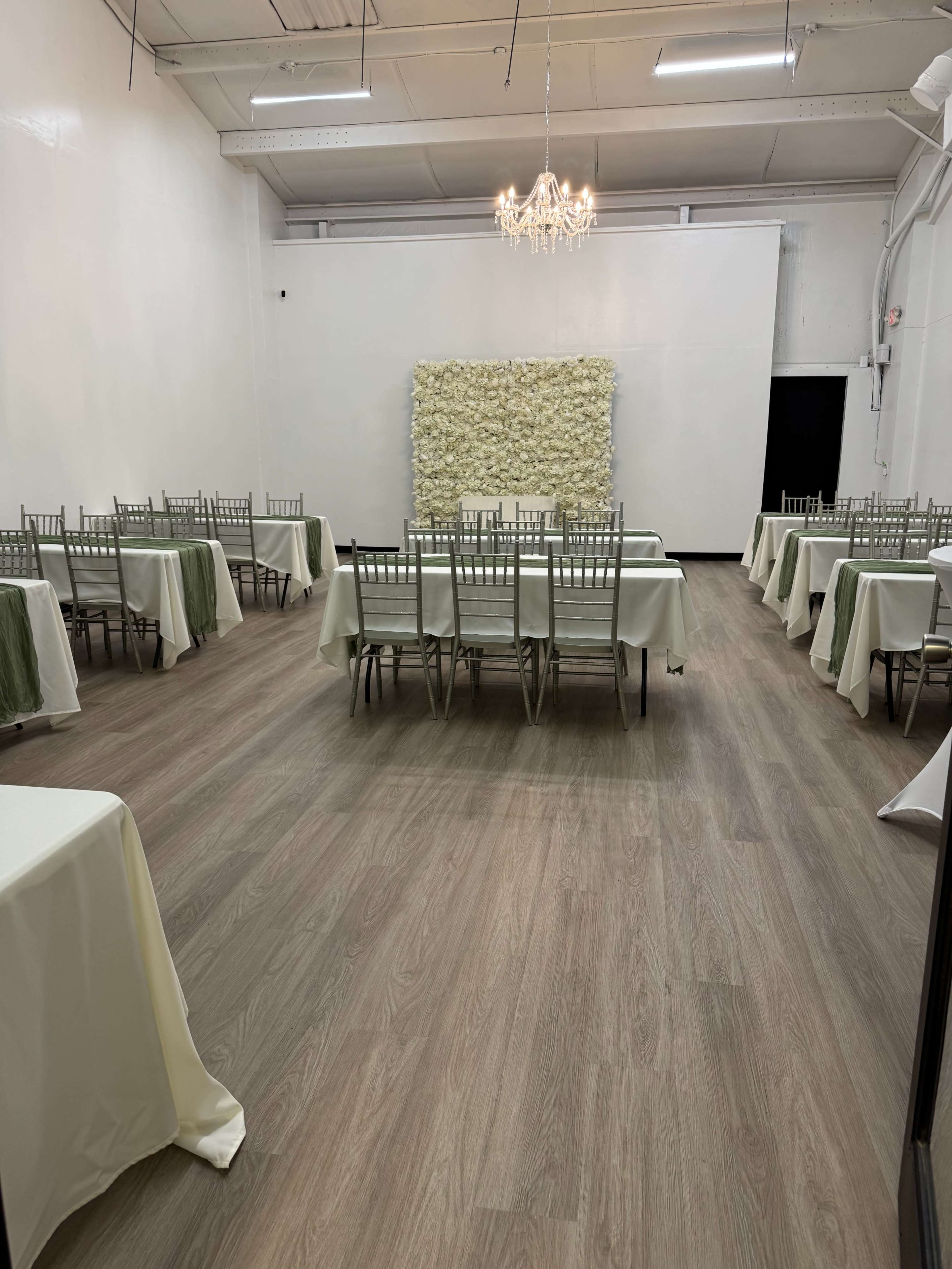 The image shows a spacious indoor room with several tables set for a gathering, featuring light-colored tablecloths and a floral wall decoration at the back.