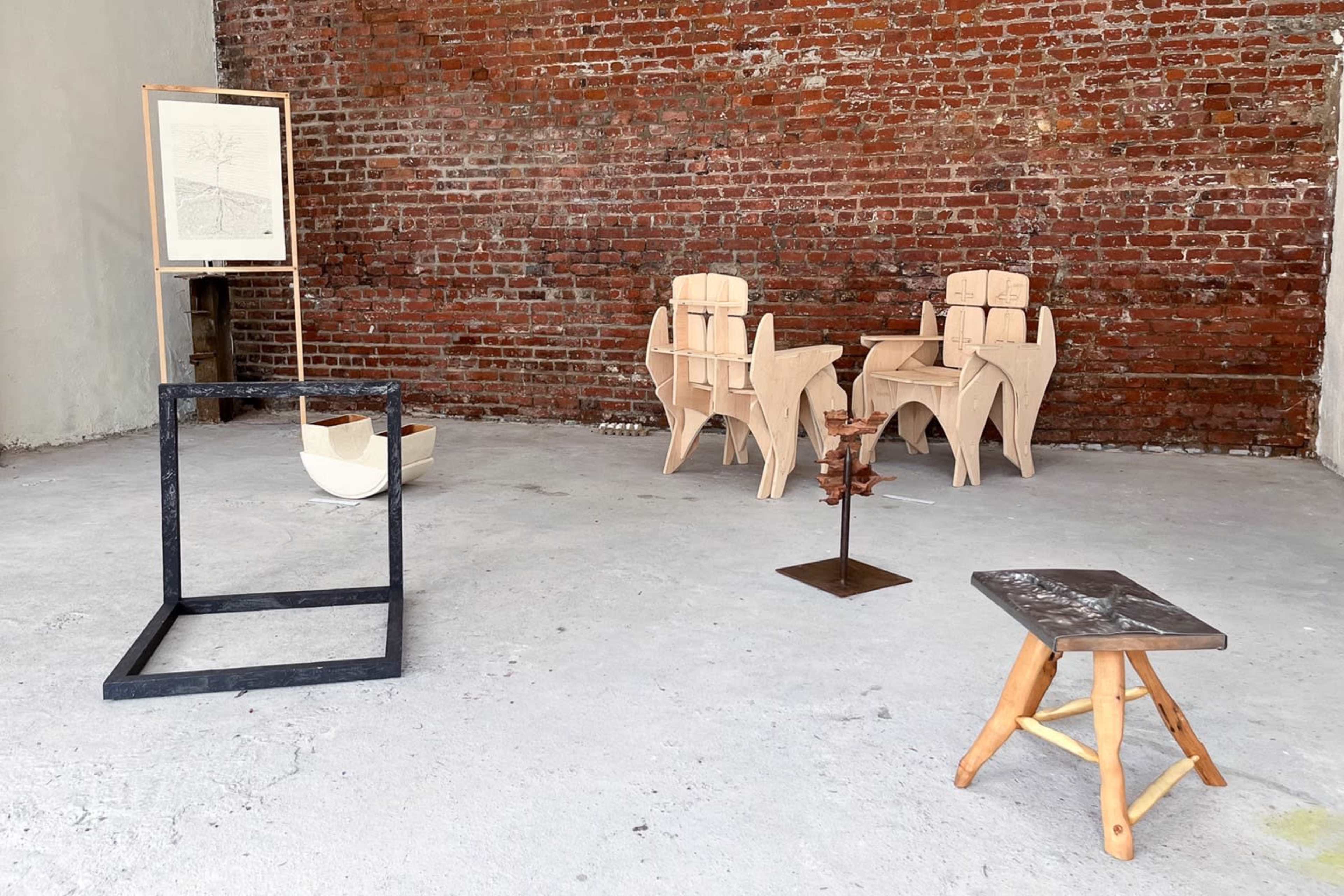An art gallery space with several wooden sculptures and furniture pieces arranged against a brick wall, including two wooden chairs, a framed artwork, and a stool.