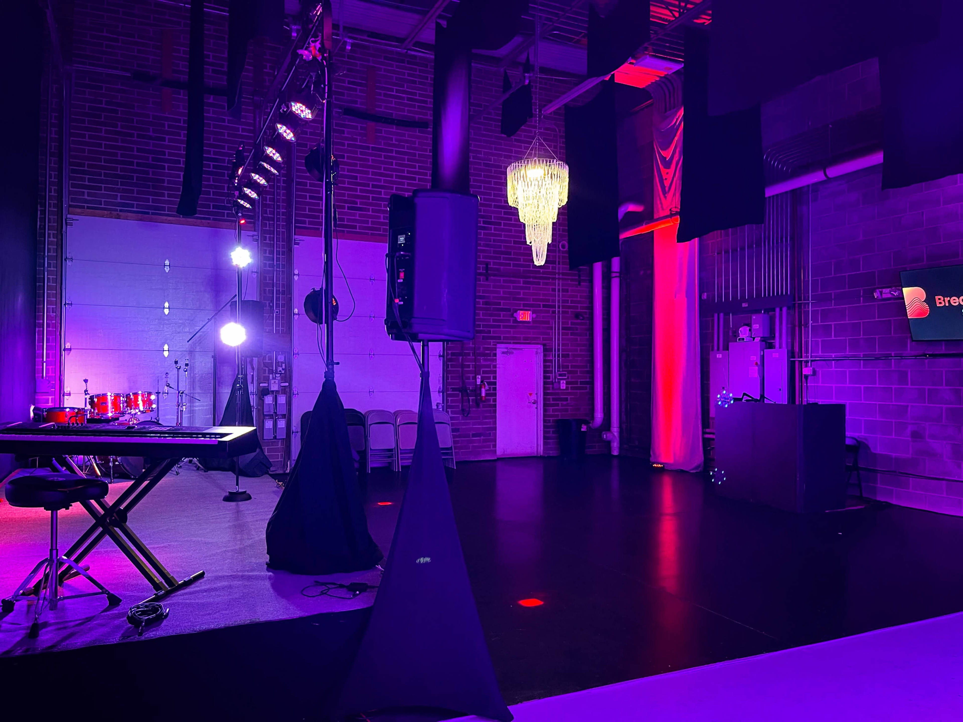 A dimly lit event space with a stage featuring a keyboard and a chandelier, surrounded by purple and red lighting.
