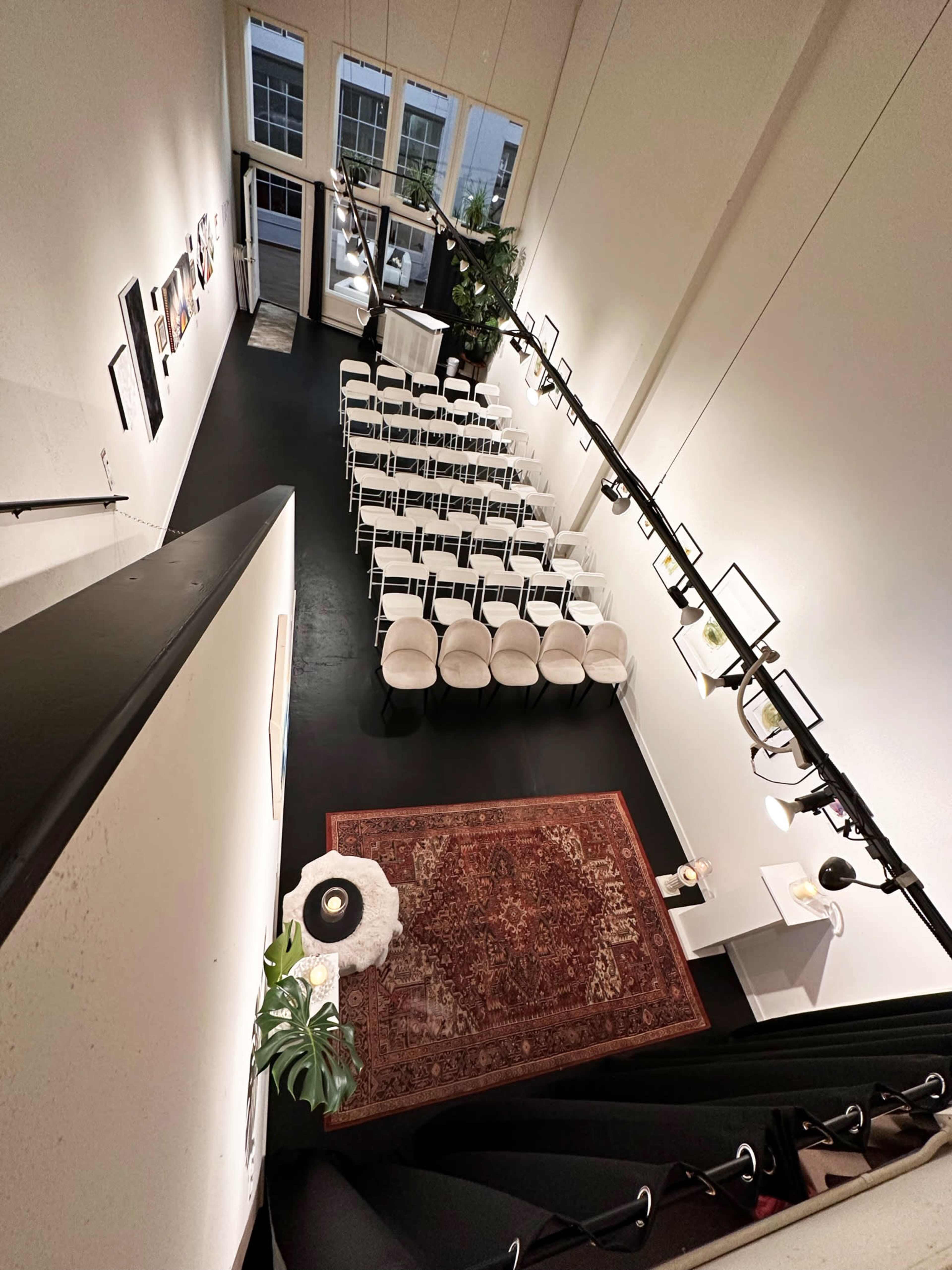 A spacious room arranged with white chairs in rows, a large rug on the floor, and decorative lighting, along with a view of large windows and greenery.