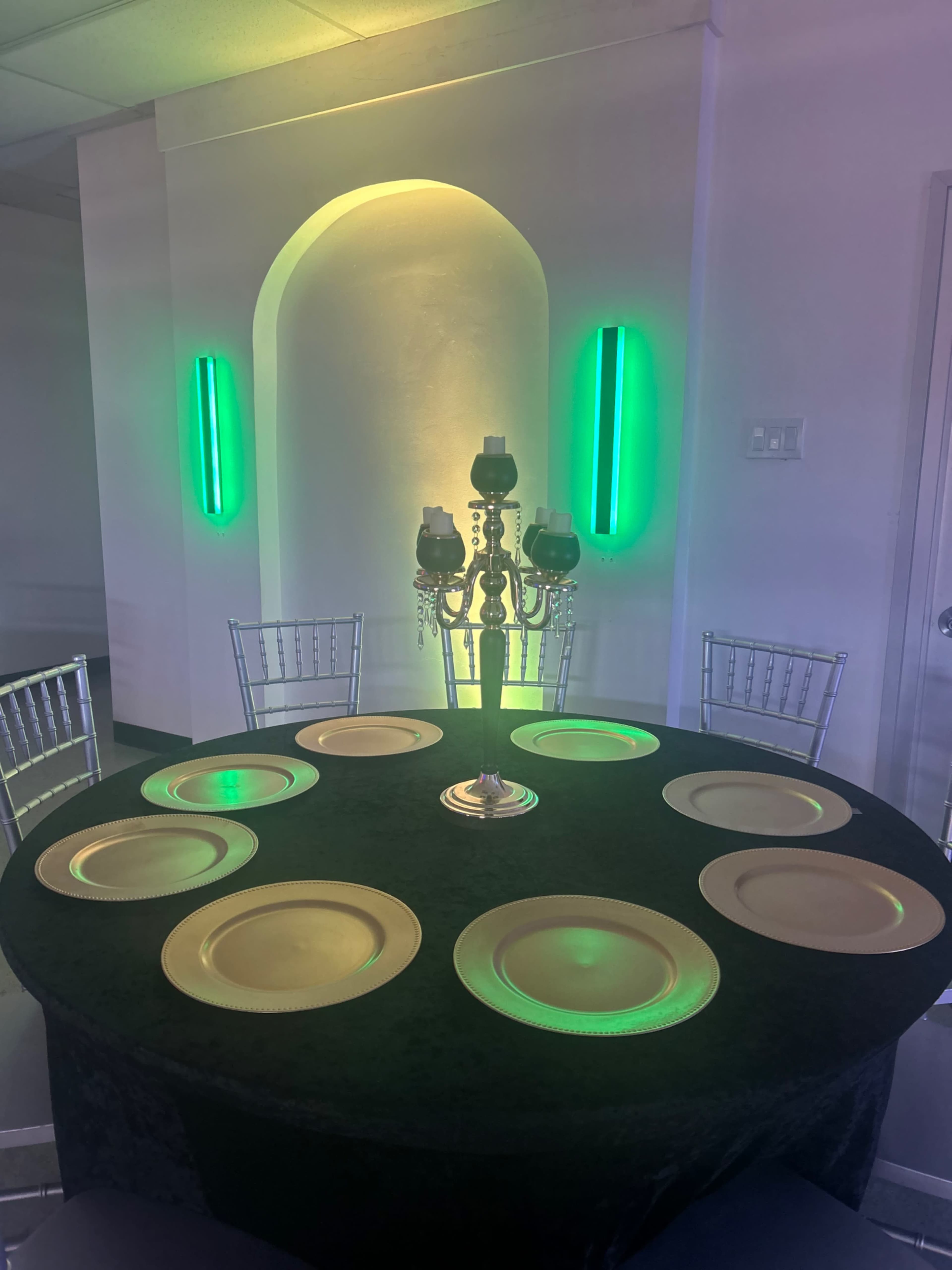 A round table is set with gold chargers and a candelabra centerpiece, surrounded by eight silver chairs in a room with green lighting.