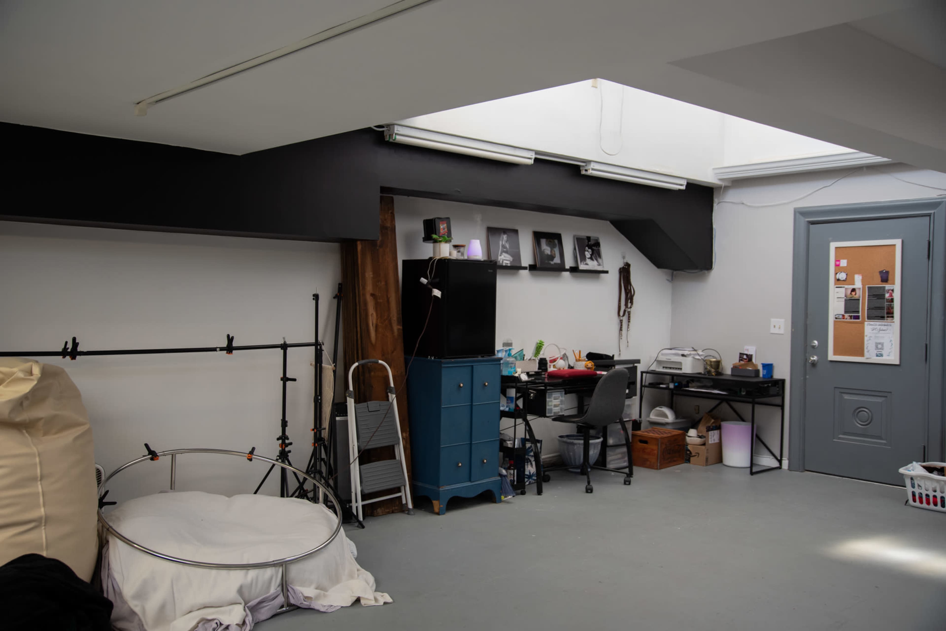 A spacious room with a blue cabinet, a desk with a computer, and a photography backdrop stand, illuminated by natural light from a skylight.