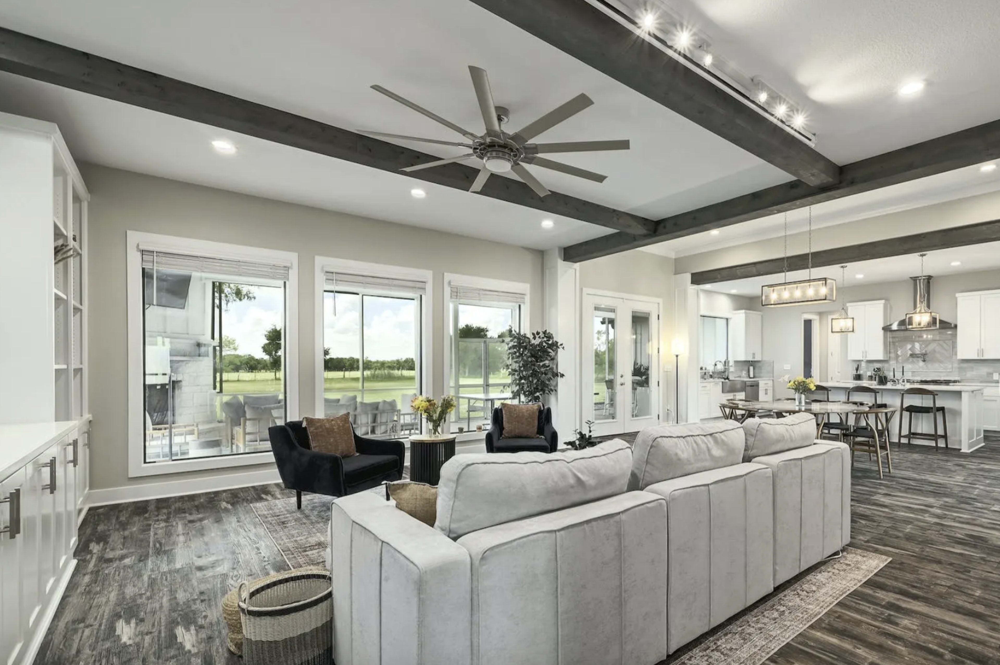 A modern living room with large windows, a white sectional sofa, and a ceiling fan, leading to a kitchen area with a dining table and bar stools.