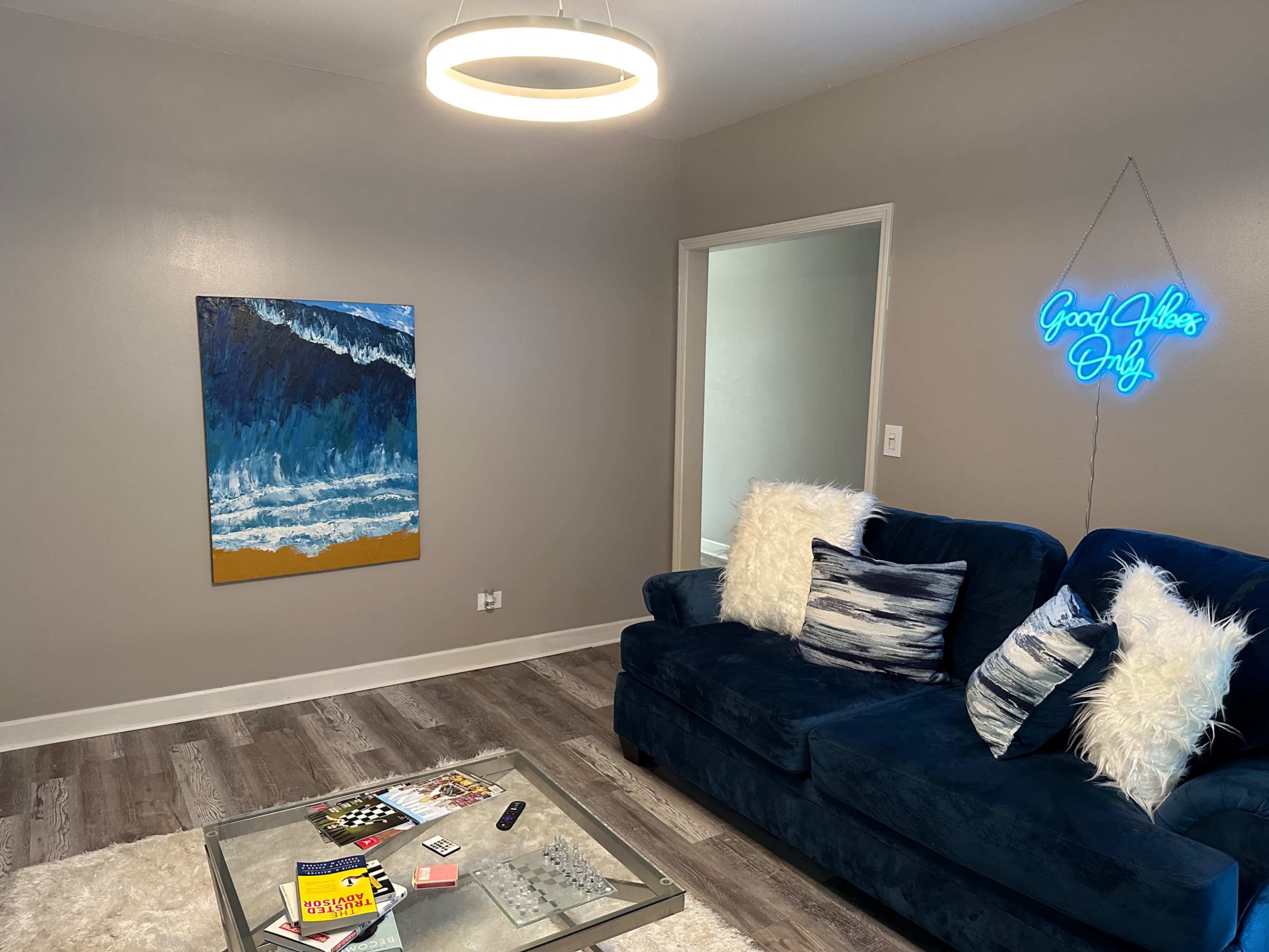 A modern living room features a blue couch with decorative pillows, a glass coffee table with books and games, a colorful ocean-themed painting, and a neon sign that reads "Good Vibes Only."