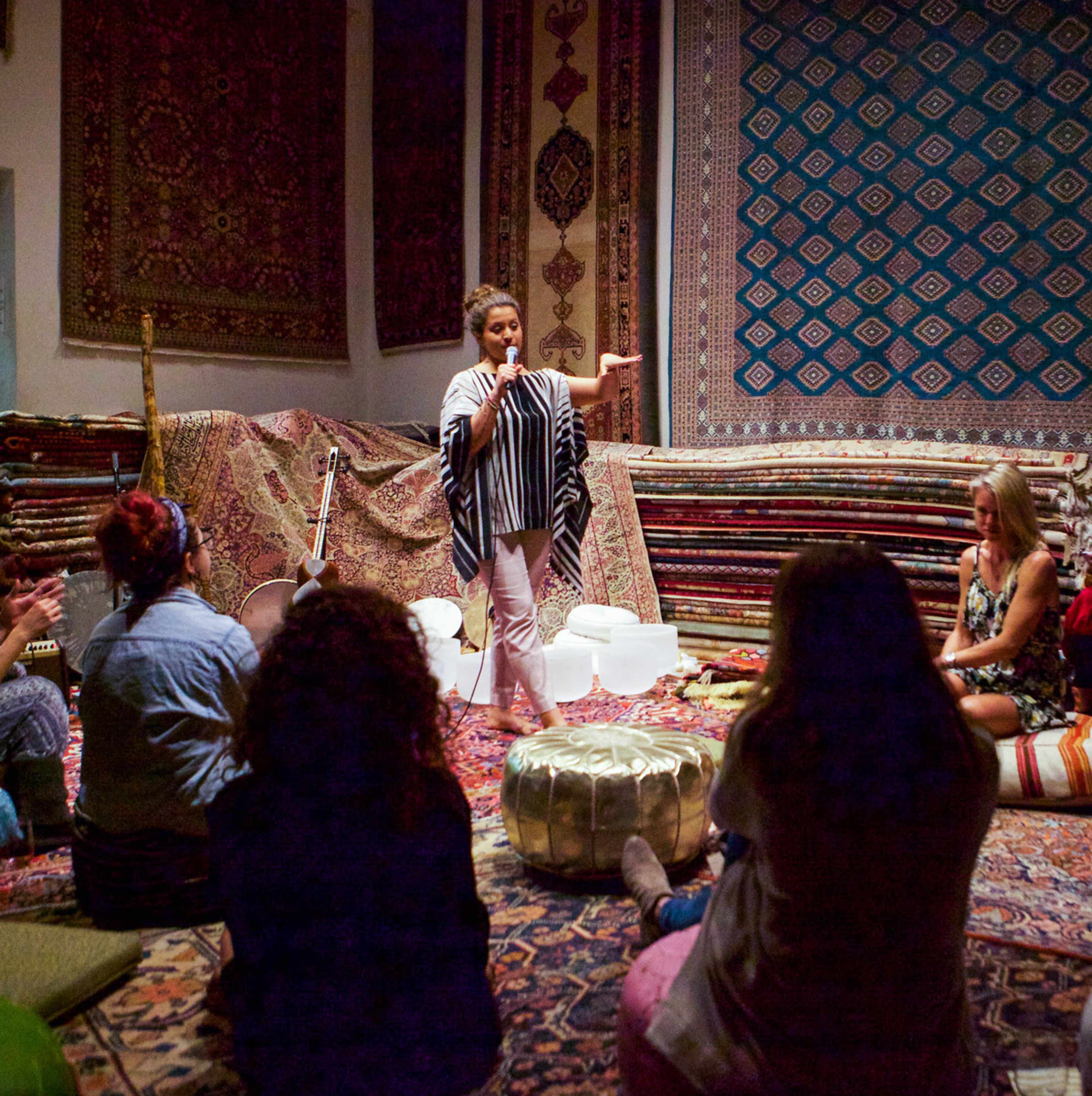 A woman speaks while standing in front of an audience seated on a patterned rug surrounded by colorful tapestries.