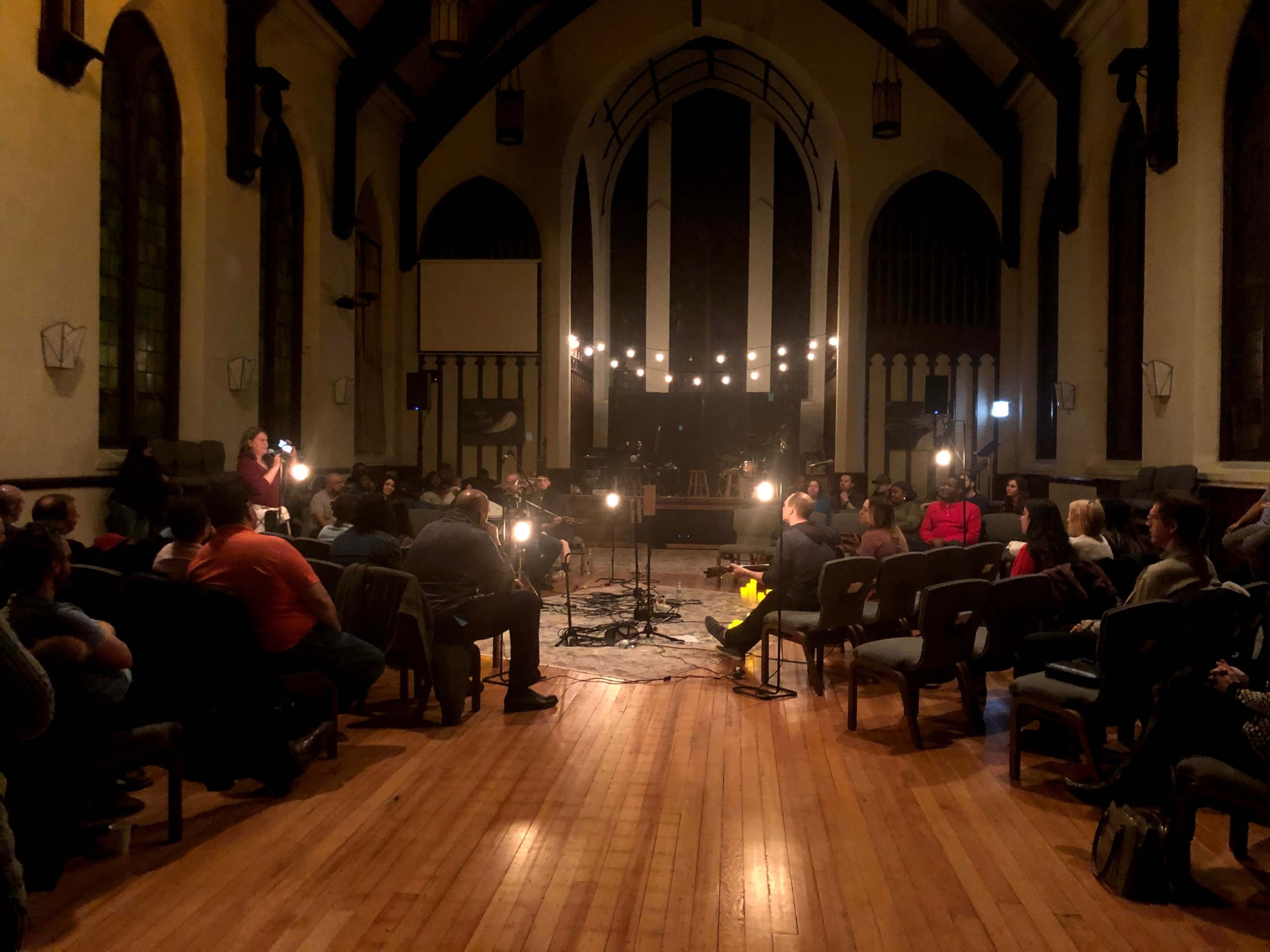 A gathering of people is seated in an indoor space with a wooden floor, surrounded by dim lighting and string lights.