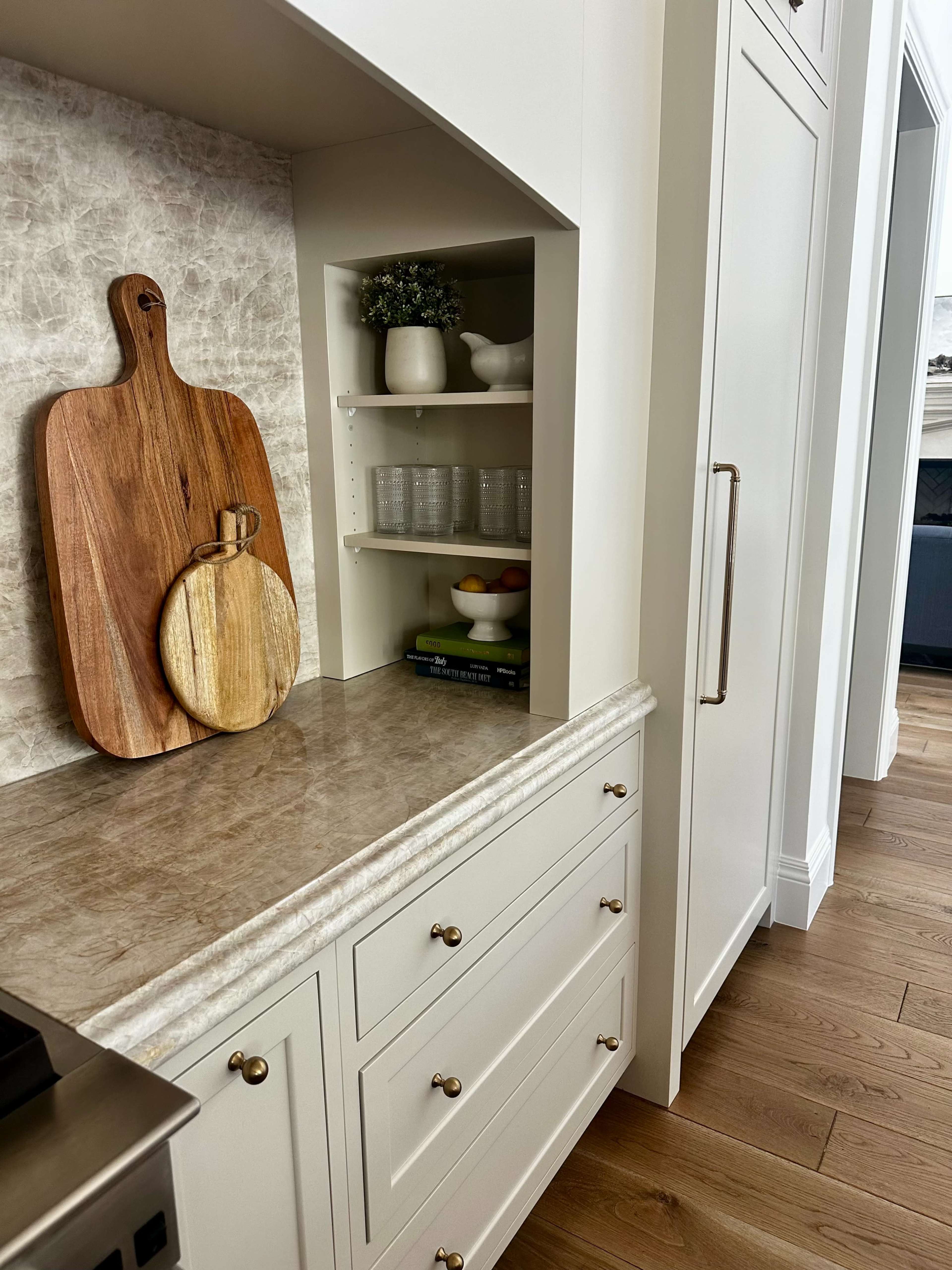 The image shows a modern kitchen area featuring a wooden cutting board, kitchen utensils, and glassware arranged on a countertop and shelves.