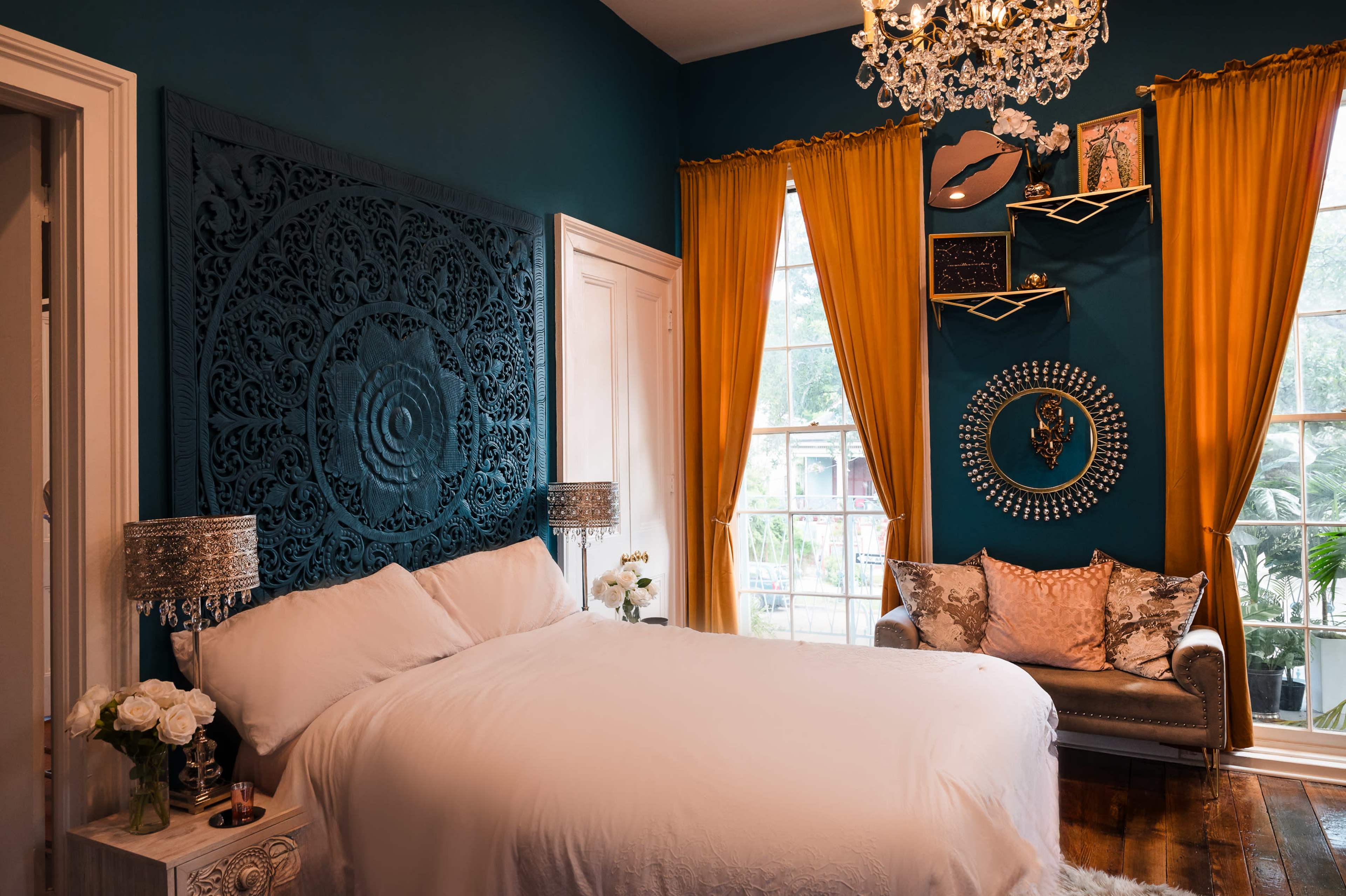 The image shows a bedroom with a large white bed, an ornate dark blue feature wall, yellow curtains, a crystal chandelier, and a cozy seating area with decorative pillows.