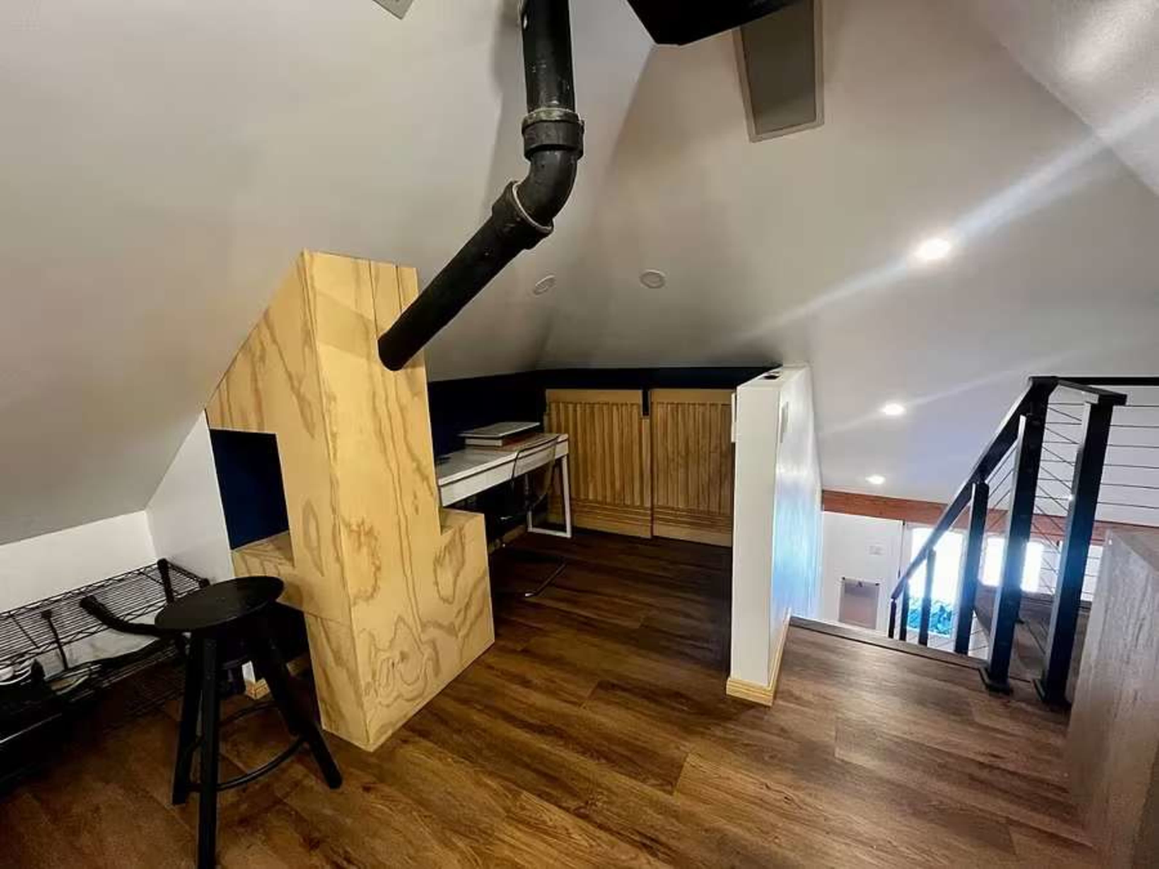 The image shows a small, modern loft space featuring wooden flooring, a kitchenette area with built-in cabinets, a black pipe, and a staircase leading to another level.