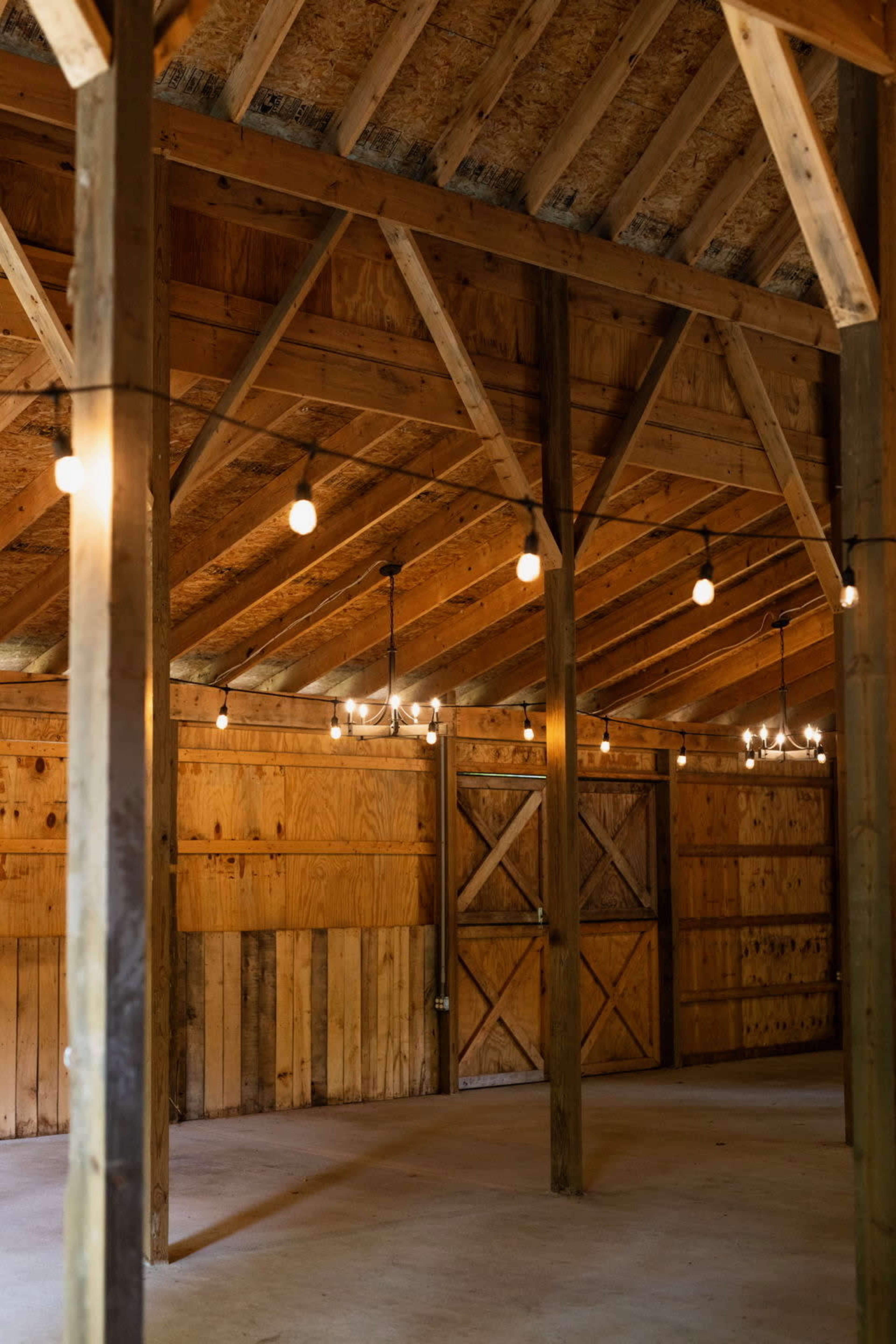 The interior of a wooden barn features exposed beams, a concrete floor, and string lights hanging from the ceiling.