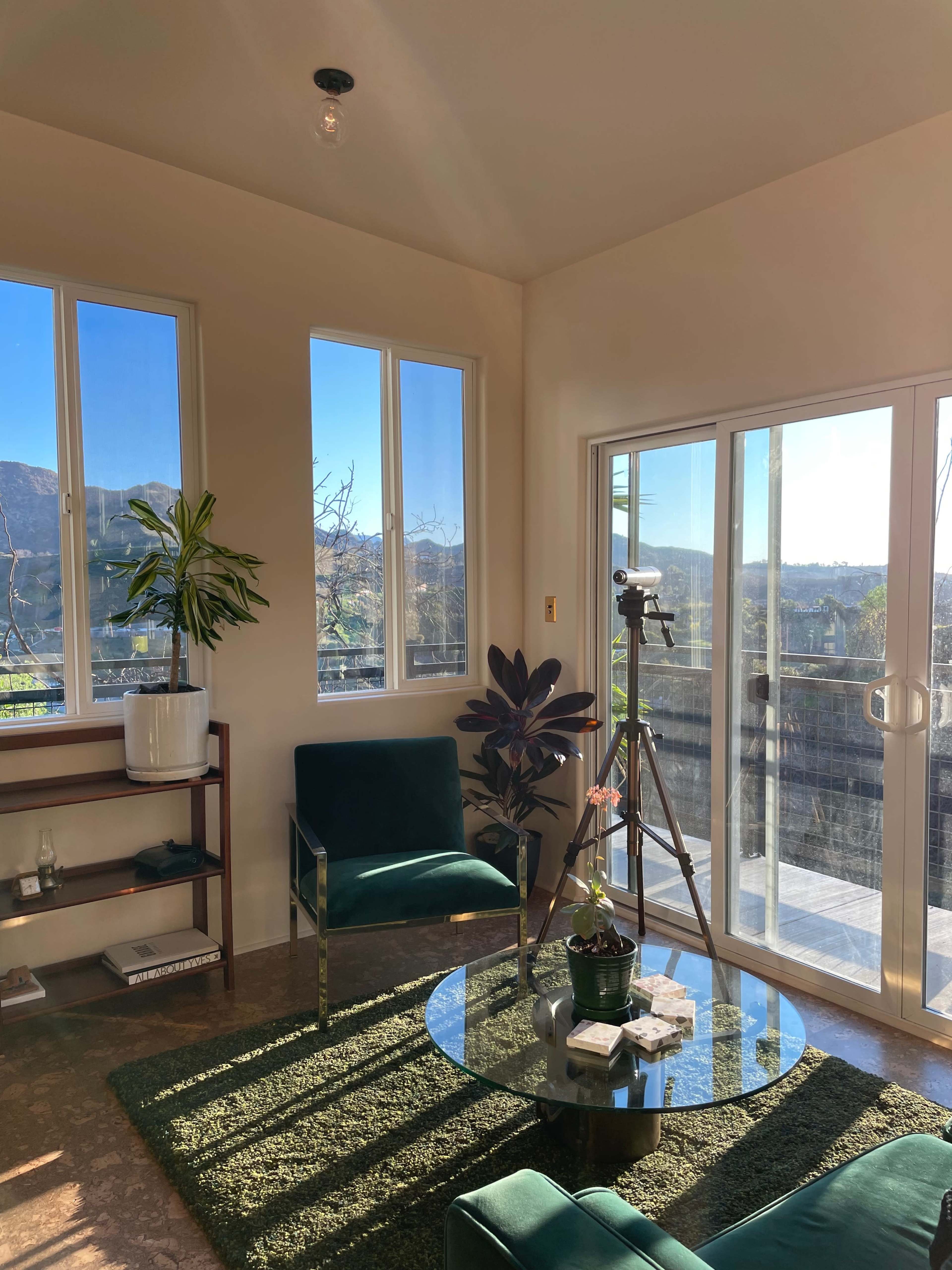 A sunlit room features large windows, green seating, a glass coffee table, and a tripod with a camera.